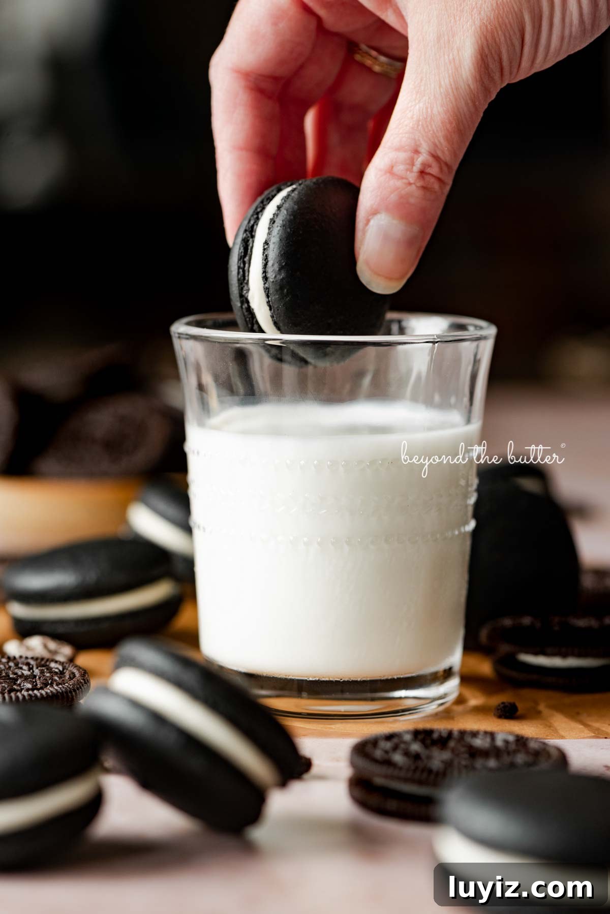 Oreo Cookie Macarons: Step-by-Step Recipe for Perfect Bites 4 Hand dipping an oreo macaron in a glass of milk with more oreo macarons and oreo cookies around it