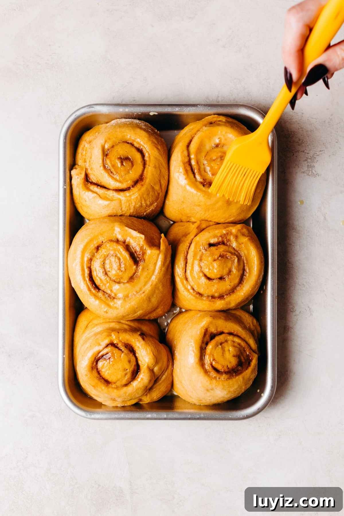 Pumpkin Cinnamon Rolls with Cream Cheese Frosting Recipe 15 Brushing egg wash onto six pumpkin cinnamon roll slices on a metal baking sheet before baking.