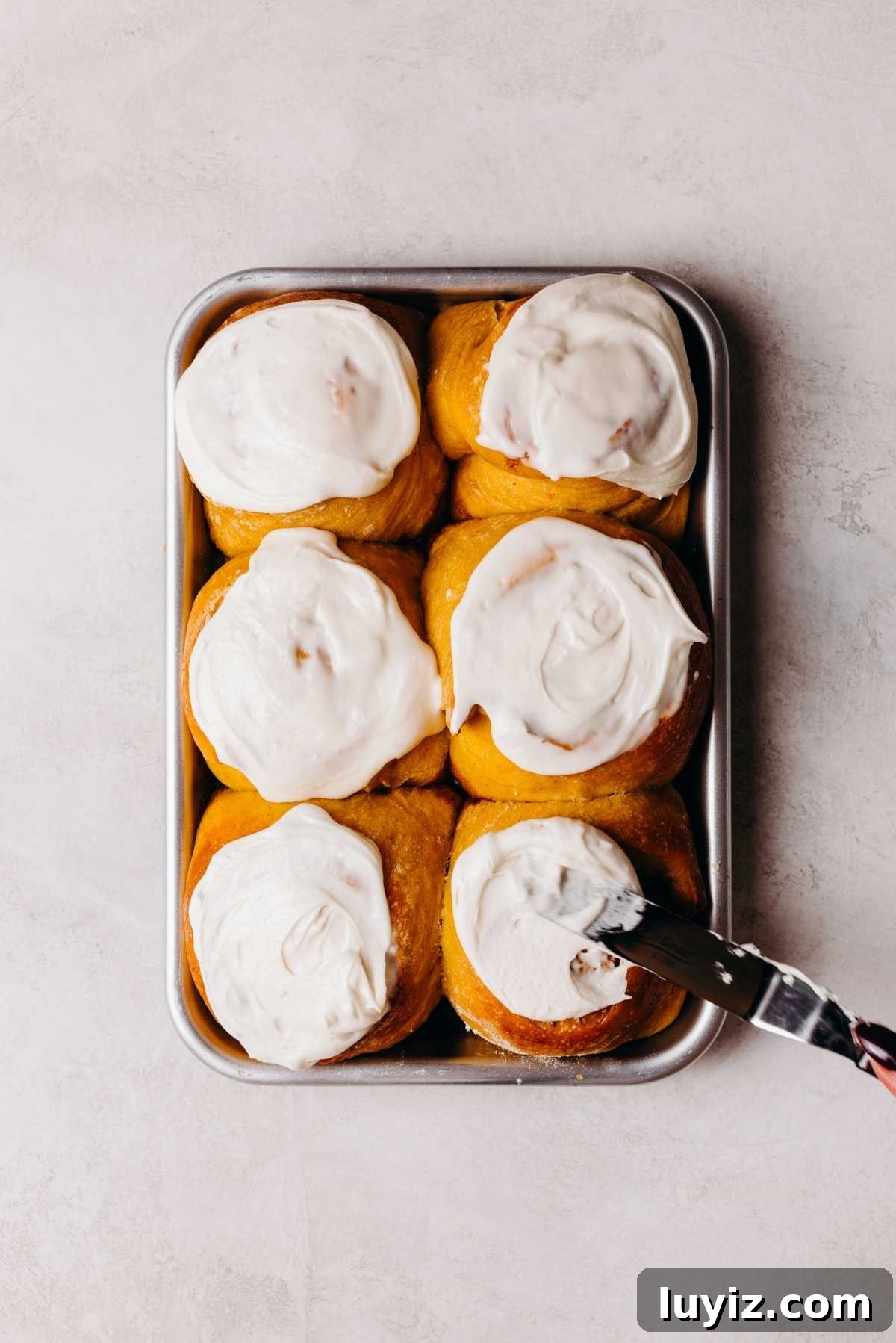 Pumpkin Cinnamon Rolls with Cream Cheese Frosting Recipe 17 Pumpkin cinnamon rolls on a metal baking sheet being topped with maple frosting using an offset spatula.