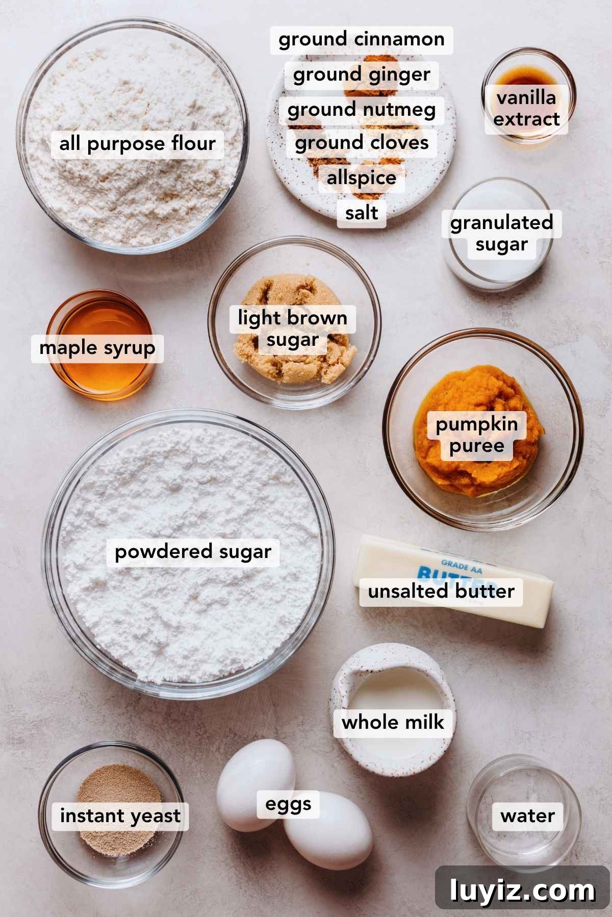 Pumpkin Cinnamon Rolls with Cream Cheese Frosting Recipe 5 Ingredients in various size bowls labeled as all purpose flour, ground cinnamon, ground ginger, ground nutmeg, ground cloves, allspice, salt, vanilla extract, maple syrup, light brown sugar, granulated sugar, pumpkin puree, powdered sugar, unsalted butter, whole milk, instant yeast, eggs, and water on a white marbled background.