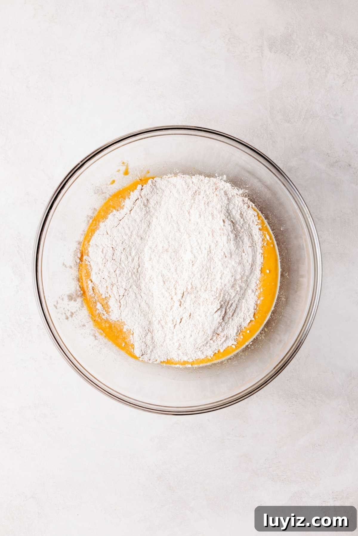Pumpkin Cinnamon Rolls with Cream Cheese Frosting Recipe 8 Flour mixture added to pumpkin cinnamon roll wet ingredients in a medium size glass mixing bowl on a white and grey marbled background.
