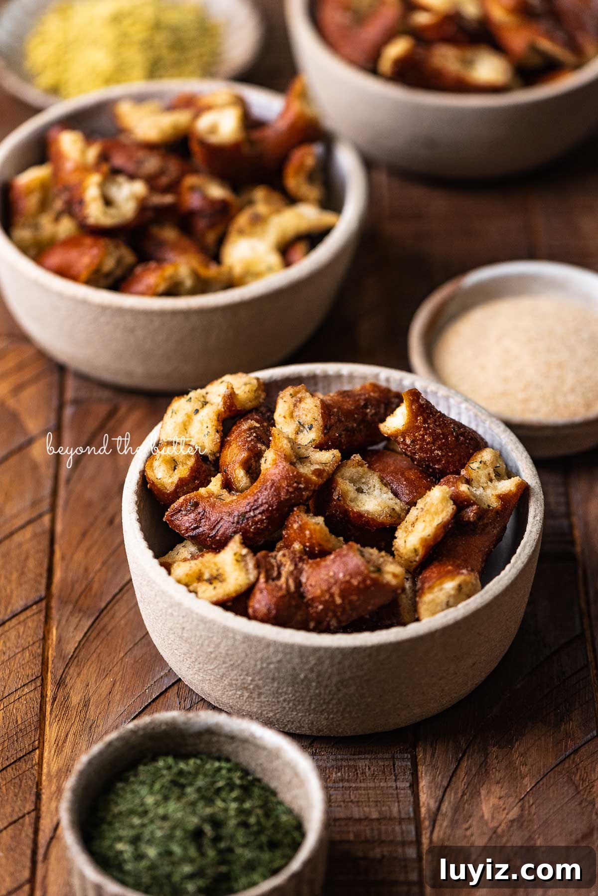 Savory Seasoned Pretzels: Flavorful Homemade Snack Recipe 6 Small snacking bowls filled with seasoned pretzels with dill weed and garlic powder in side bowl on dark wood background from BeyondtheButter.com | © Beyond the Butter