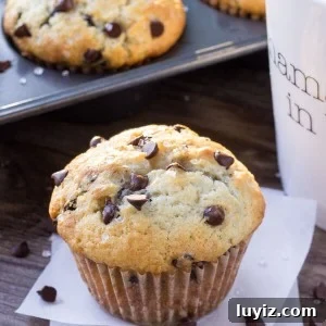 One big, fluffy bakery-style chocolate chip muffin with a cup of coffee and pan full of muffins in the background.