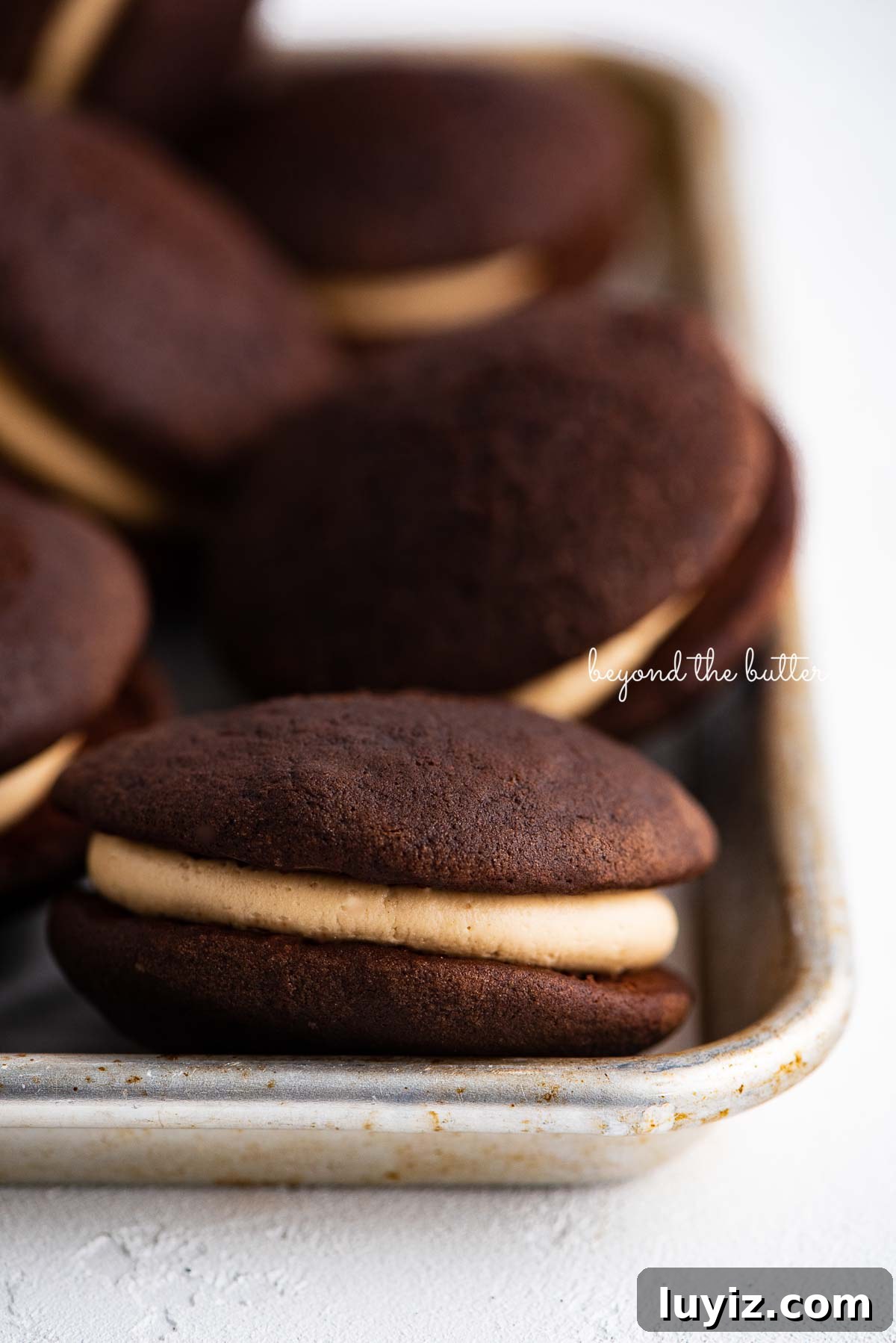 Chocolate Peanut Butter Whoopie Pies Recipe — Soft, Gooey Sandwich Cookies 3 Chocolate peanut butter whoopie pies on a parchment lined baking pan | © Beyond the Butter®