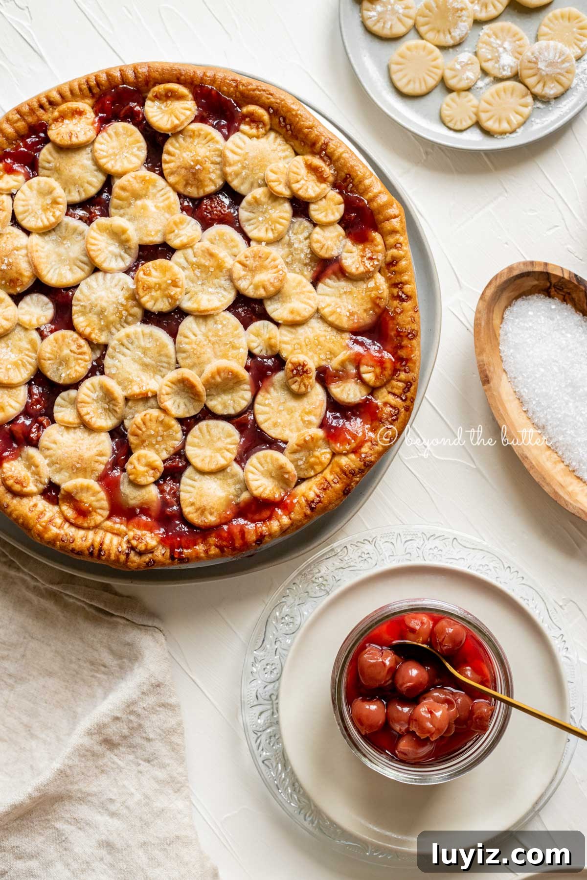 Classic Homemade Cherry Pie Recipe for Perfect Crust and Filling 3 Overhead image of cherry pie surrounded by plates, jar of cherries, sparkling sugar, and pie crust toppers | All Images © Beyond the Butter™