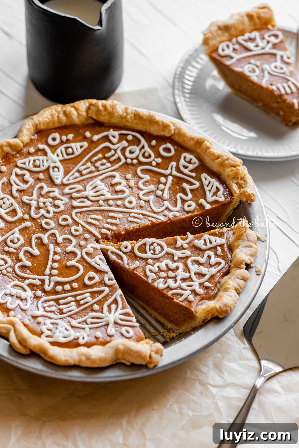 Perfect Pumpkin Pie Recipe for Creamy, Flaky Crust 6 Angled image of cut pumpkin pie with a piped cream cheese frosting decoration surrounded by pie server, cinnamon sticks, plates, and small pitcher of milk