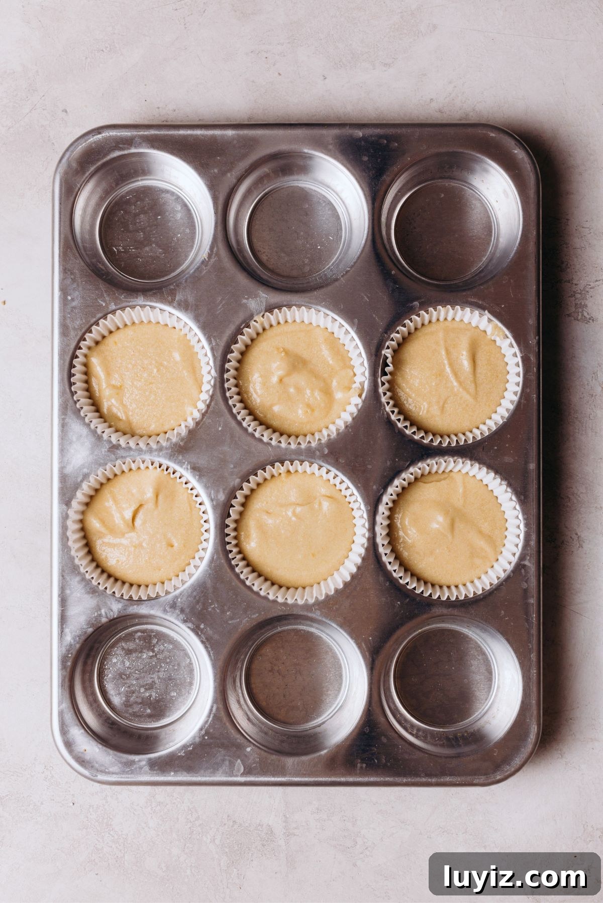 Chocolate Chip Cookie Dough Cupcakes Recipe with Gooey Centers 12 Metal cupcake tin with six filled cavities with vanilla cupcake batter on a white marbled background.