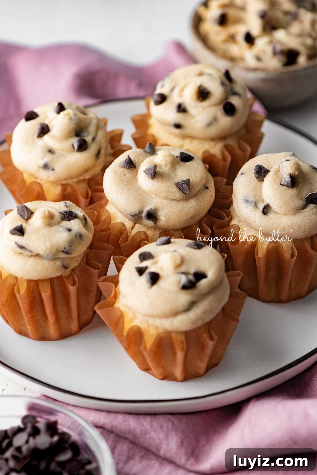 Chocolate Chip Cookie Dough Cupcakes Recipe with Gooey Centers 20 Black and white ceramic dessert plate placed on a pink cloth napkin with six chocolate chip cookie dough cupcakes.