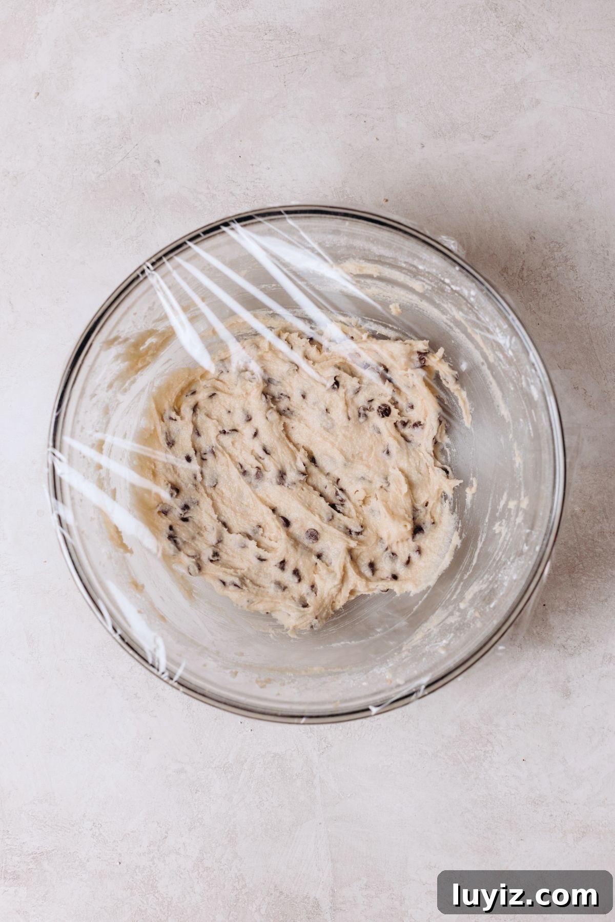 Chocolate Chip Cookie Dough Cupcakes Recipe with Gooey Centers 9 Glass mixing bowl with edible chocolate chip cookie dough covered in plastic wrap on a white marbled background.