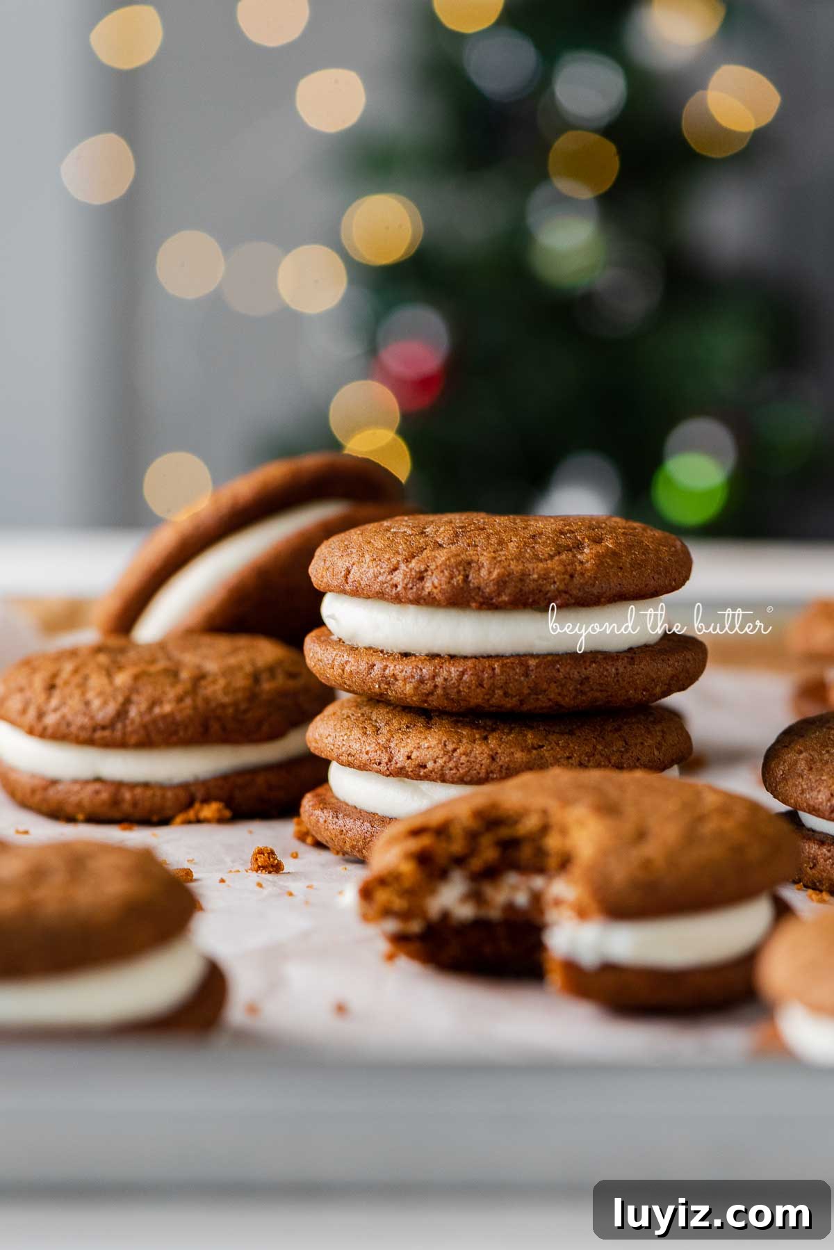 Gingerbread Whoopie Pies Recipe: Soft Molasses Sandwich Cookies 5 Stacked gingerbread whoopie pies on white parchment paper with holiday tree background | © Beyond the Butter®