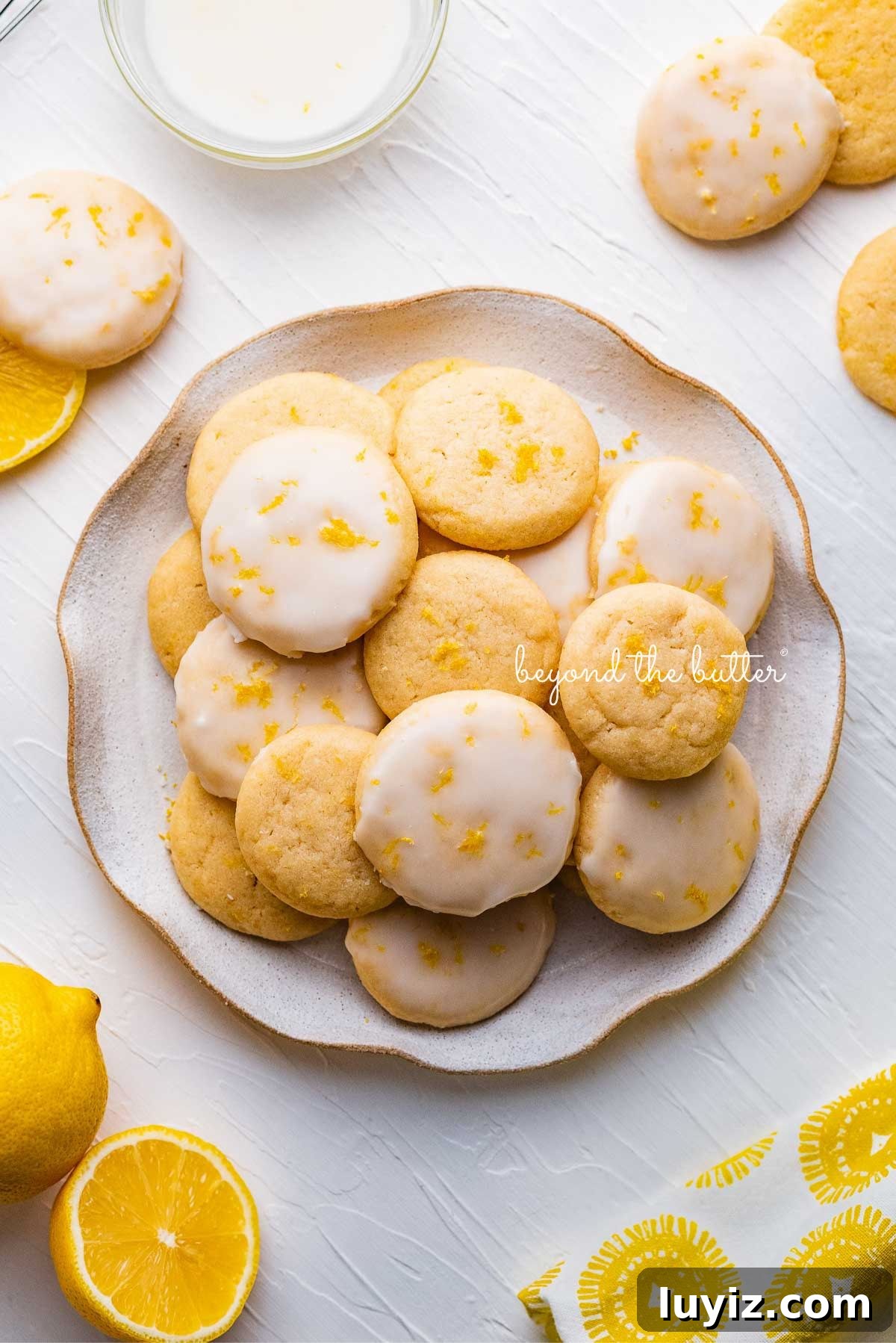 Zesty Lemon Shortbread Cookies: Crispy, Buttery Citrus Treats 14 Plate full of lemon shortbread cookies surrounded by a lemon themed cloth napkin, lemons, and glass of milk on a white background.