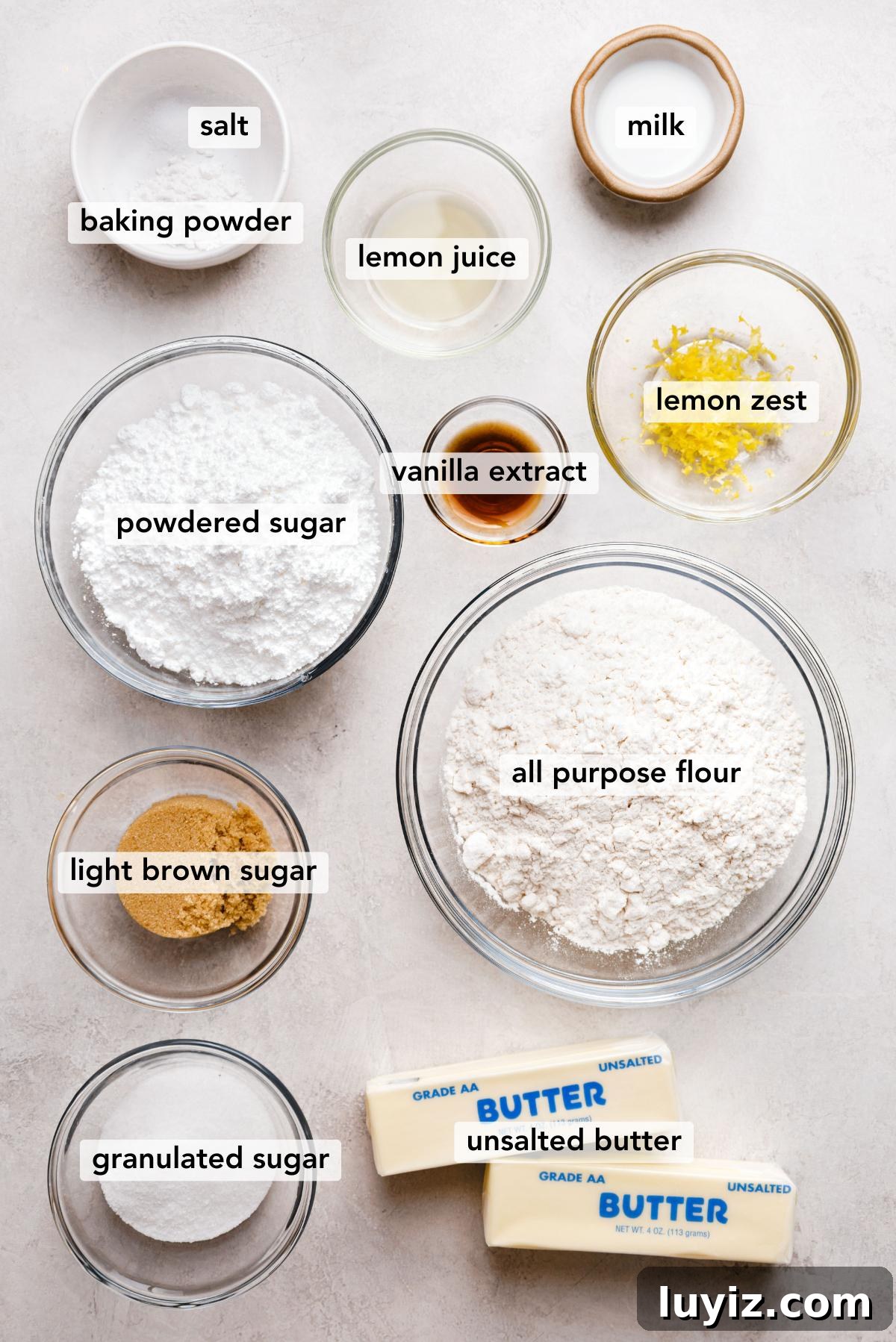 Zesty Lemon Shortbread Cookies: Crispy, Buttery Citrus Treats 5 Ingredients in various size bowls labeled as salt, baking powder, milk, powdered sugar, vanilla extract, lemon juice, lemon zest, light brown sugar, all purpose flour, granulated sugar, and unsalted butter on a white marbled background.