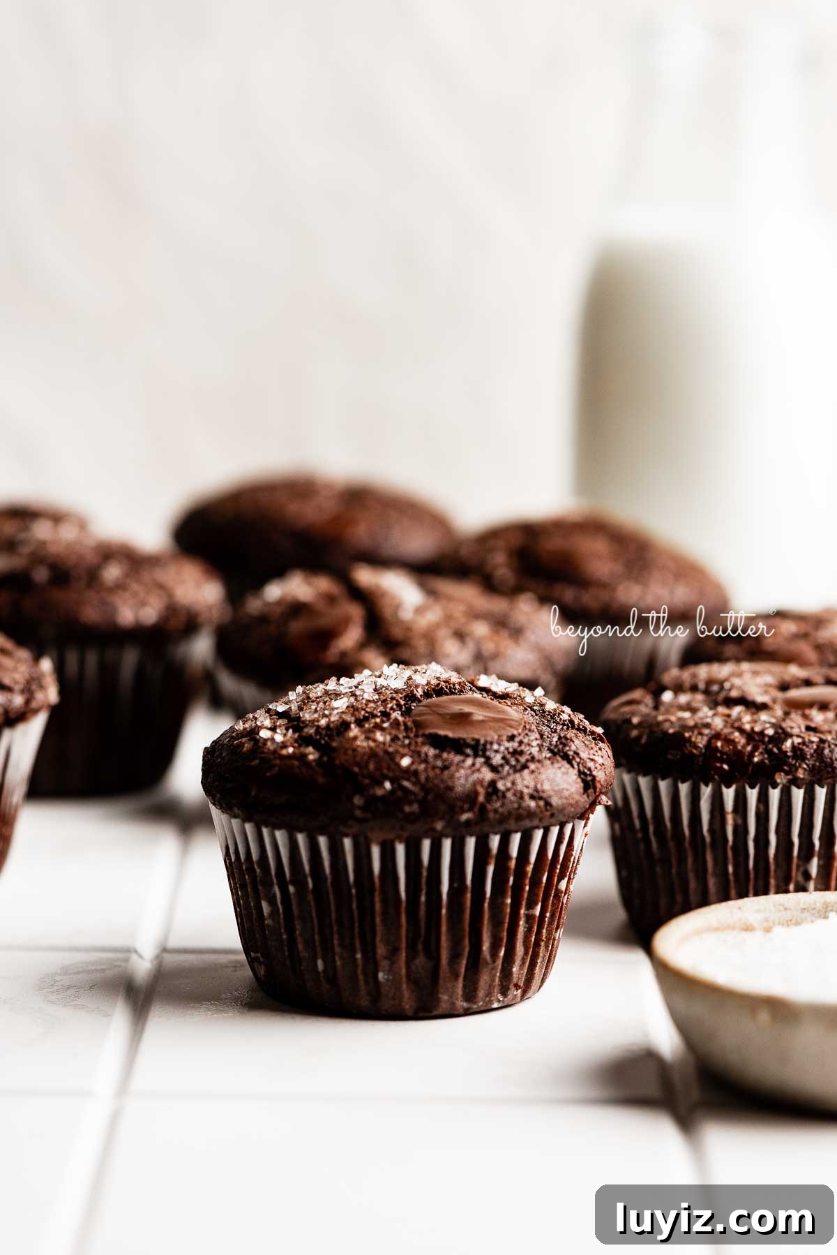 Ultimate Double Chocolate Muffins Recipe – Moist & Fudgy 3 Double chocolate muffins on white tiled background with bottle of milk and small bowl of sparklin sugar to the side | © Beyond the Butter®