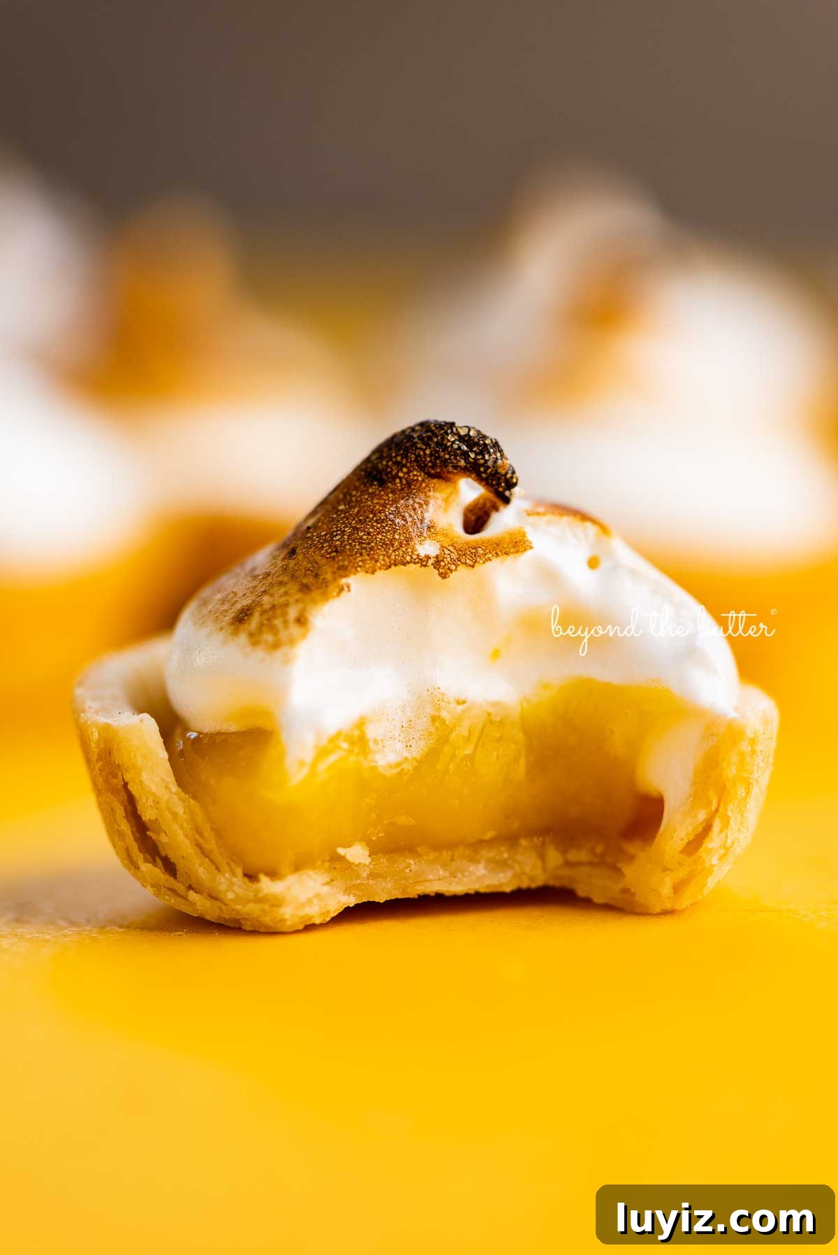 Mini Lemon Meringue Tartlets: Light, Zesty Dessert Recipe 3 Mini lemon meringue pie with bite taken out surrounded by other mini pies on a yellow background | © Beyond the Butter@
