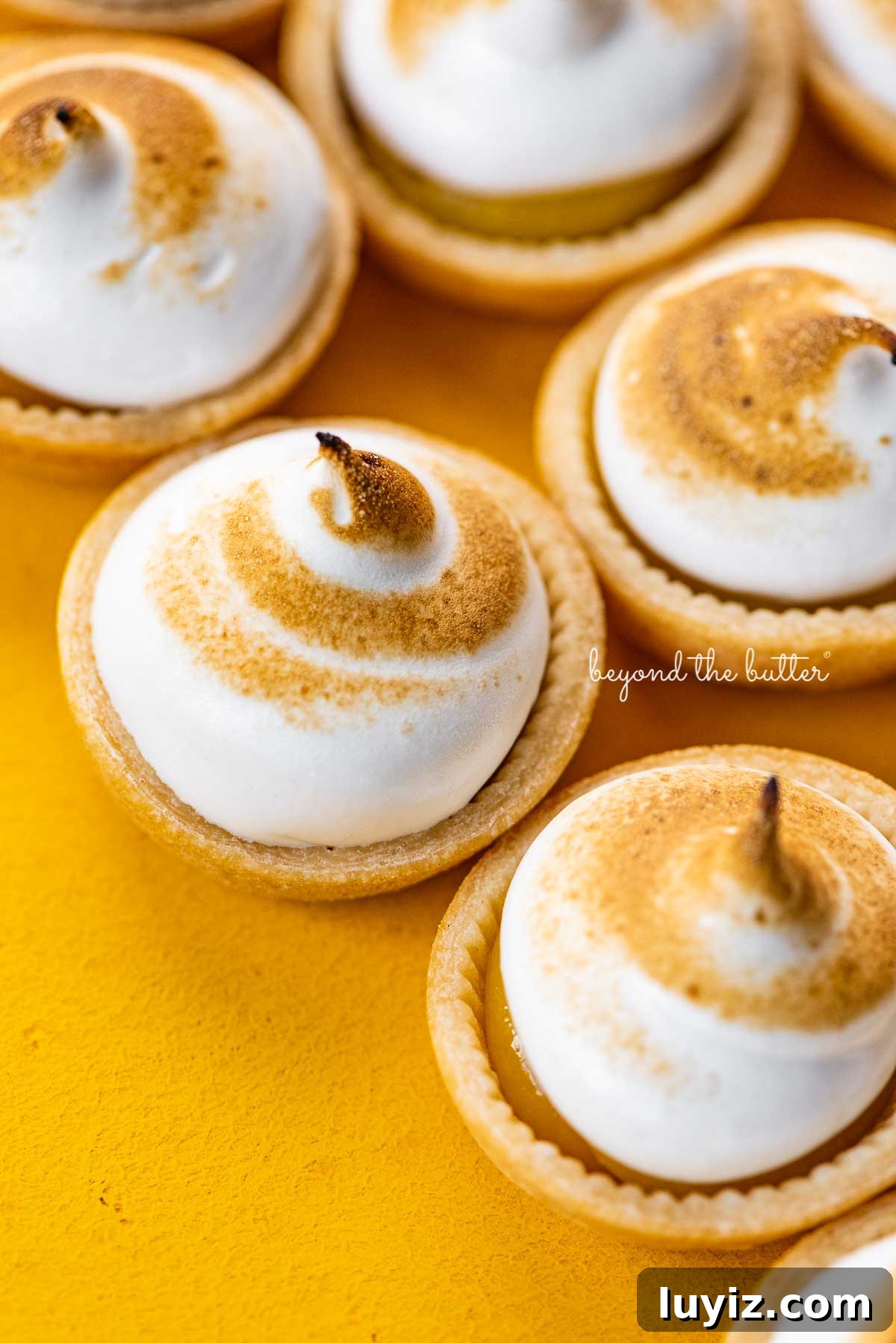 Mini Lemon Meringue Tartlets: Light, Zesty Dessert Recipe 6 Angled image of mini lemon meringue pies on a yellow background | © Beyond the Butter®