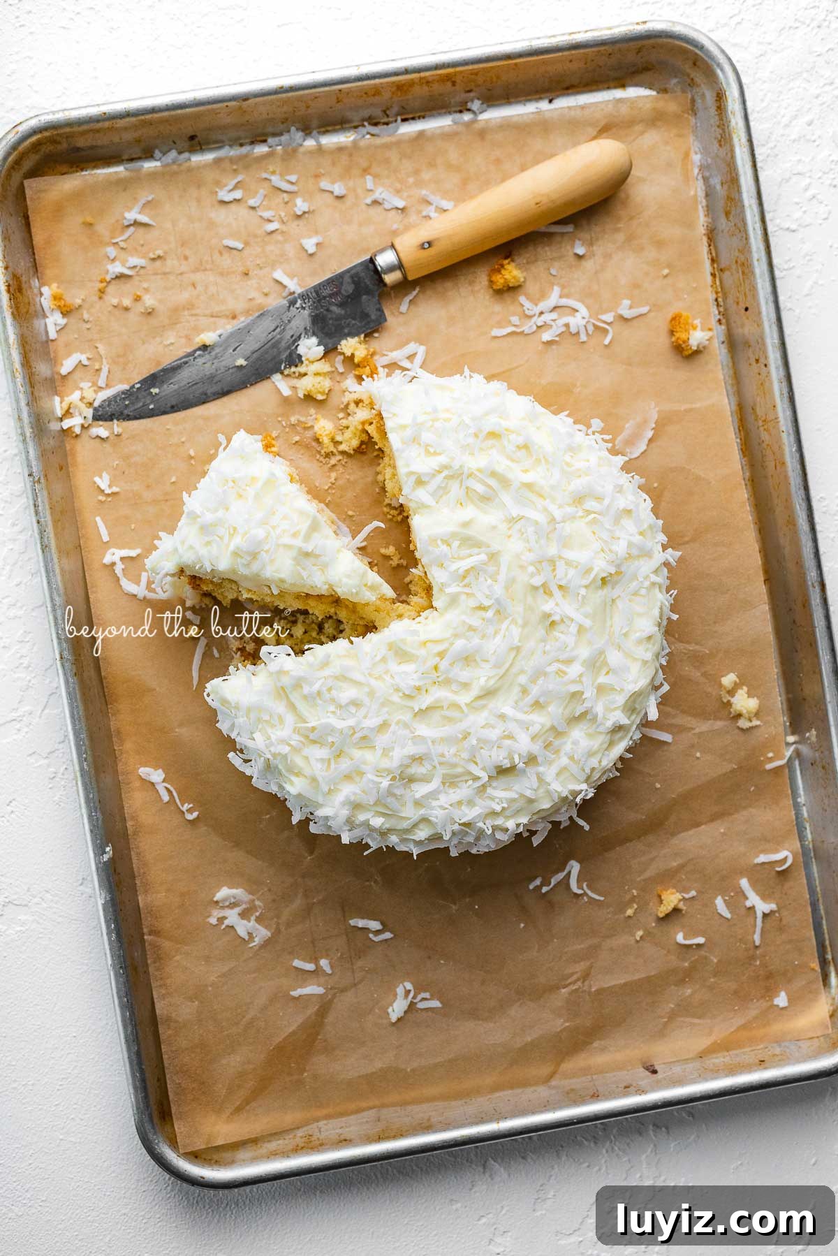 Moist Single-Layer Coconut Cake Recipe for Any Occasion 5 Single layer coconut cake frosted with coconut buttercream with slice cut out on natural parchment paper-lined baking sheet | © Beyond the Butter®
