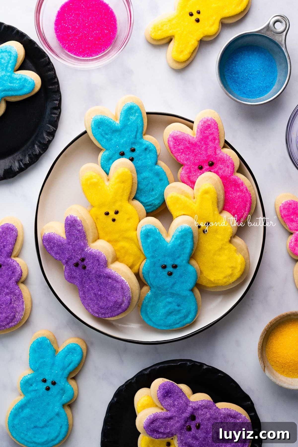 Easter Bunny Cut-Out Sugar Cookie Recipe 11 Plate full of Easter bunny cut out sugar cookies decorated in purple, pink, blue, and yellow sanding sugars with chocolate dots for eyes and noses on white marbled background.