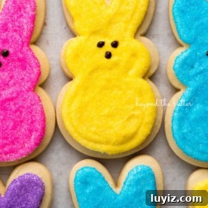 Easter Bunny Cut-Out Sugar Cookie Recipe 16 Easter bunny cut out sugar cookies topped with frosting and colors of pink, yellow, blue, and purple sanding sugars on a parchment paper lined baking sheet.