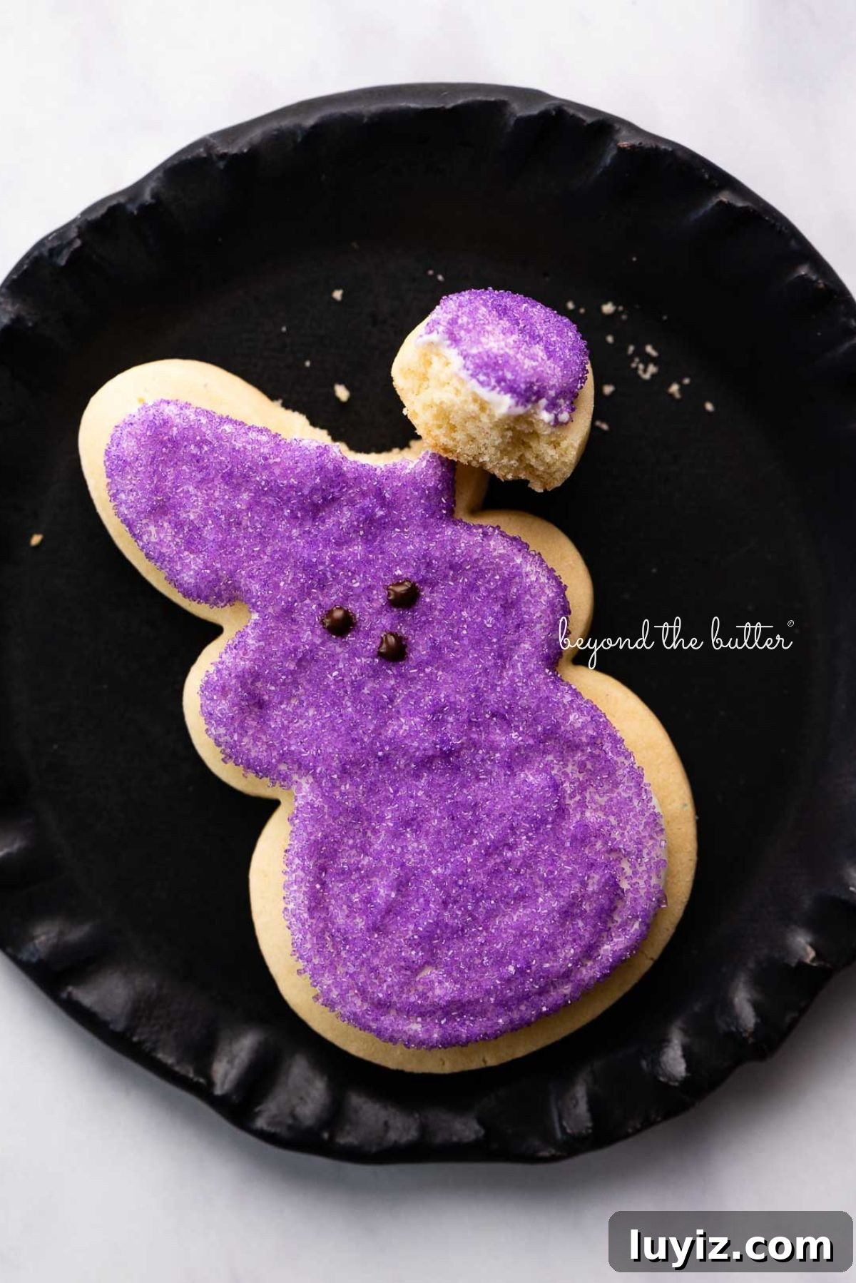 Easter Bunny Cut-Out Sugar Cookie Recipe 3 Easter bunny cut out sugar cookie decorated with purple sanding sugar and a chocolate nose and eyes with its right ear broken off on a black dessert plate.