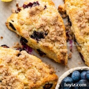 Lemon Blueberry Streusel Scones Recipe for Flaky, Berry-Packed Pastries 15 Lemon blueberry streusel scones on a parchment lined baking sheet placed next to a lemon wedge and small ceramic bowl of blueberries.