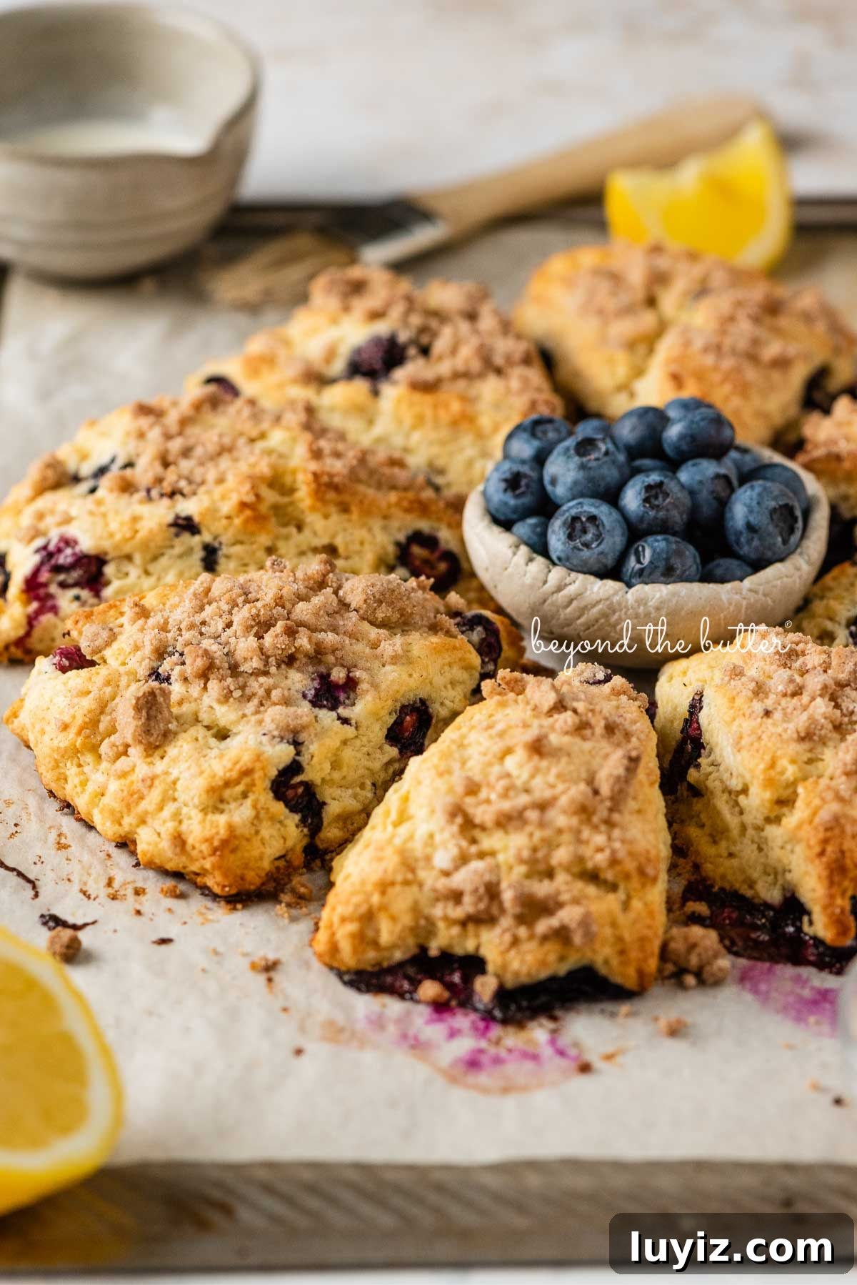 Lemon Blueberry Streusel Scones Recipe for Flaky, Berry-Packed Pastries 3 Lemon blueberry streusel scones on a parchment paper lined baking sheet placed around a small ceramic bowl of blueberries with a small bowl of milk, pastry brush, and lemon wedges around them.