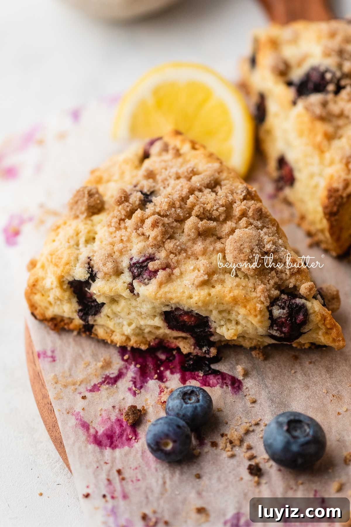 Lemon Blueberry Streusel Scones Recipe for Flaky, Berry-Packed Pastries 10 Lemon blueberry scones with streusel topping placed on a parchment paper lined round wooden serving plate with lemon wedges and blueberries placed around it.