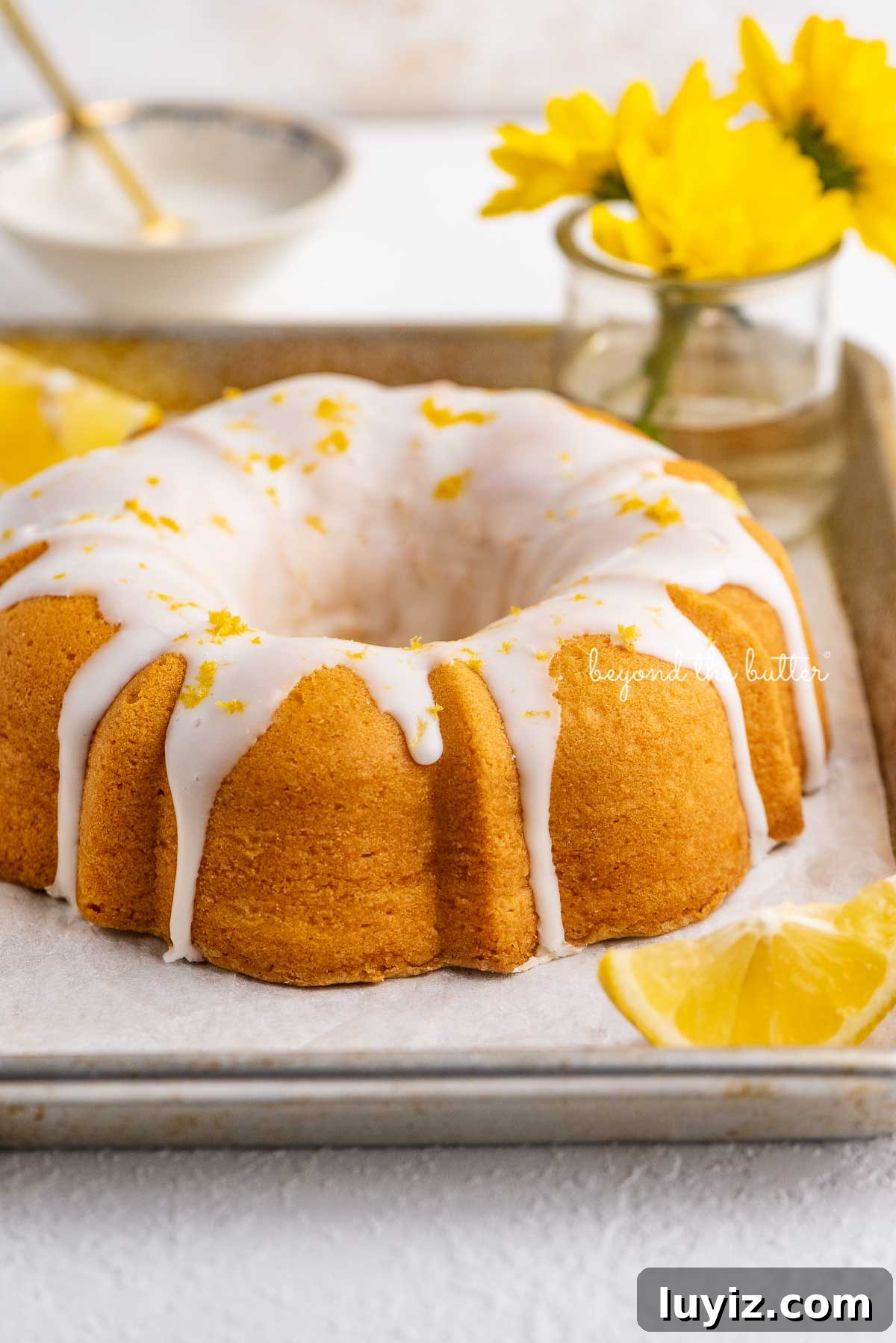 Moist Lemon Cream Cheese Pound Cake Recipe 3 Small lemon cream cheese pound cake topped with a simple lemon glaze on a parchment paper lined baking sheet with sliced lemons and small bouquet of daisies | © Beyond the Butter®
