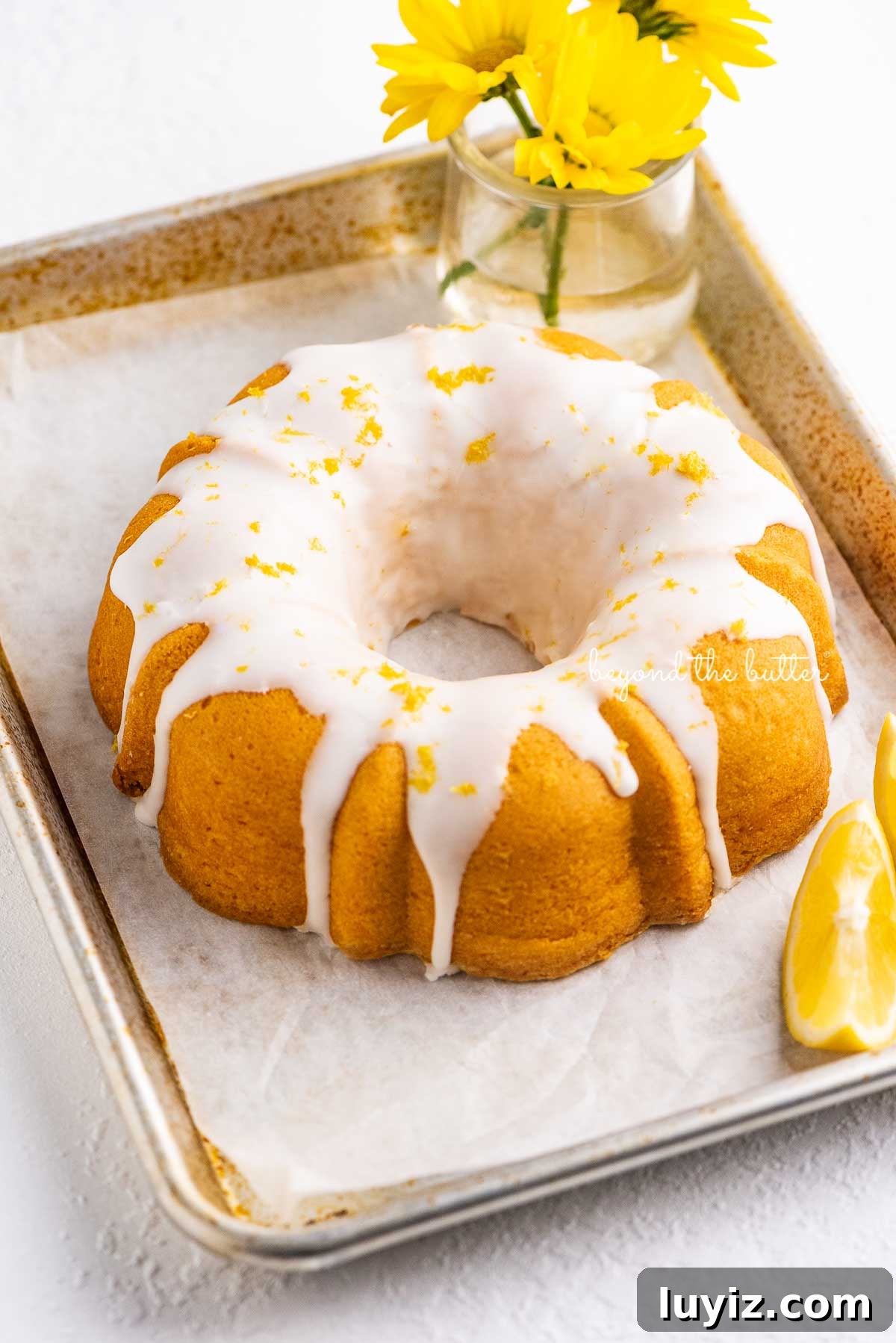 Moist Lemon Cream Cheese Pound Cake Recipe 5 Small lemon cream cheese pound cake topped with a simple lemon glaze on a parchment paper lined baking sheet with sliced lemons and small bouquet of daisies | © Beyond the Butter®