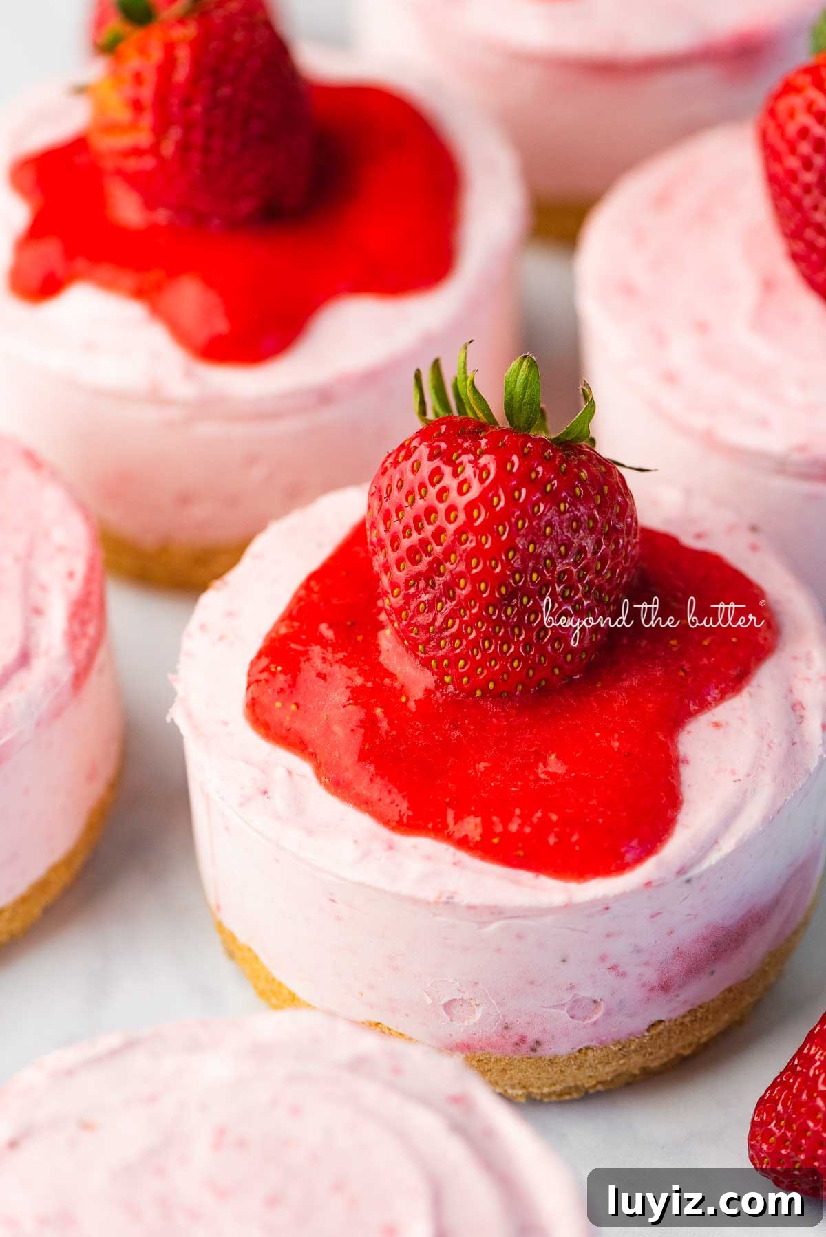 Mini No-Bake Strawberry Cheesecakes (Individual Cups) 2 Individual no-bake strawberry cheesecakes topped with strawberry glaze and a fresh strawberry on a parchment paper-lined baking sheet | © Beyond the Butter®