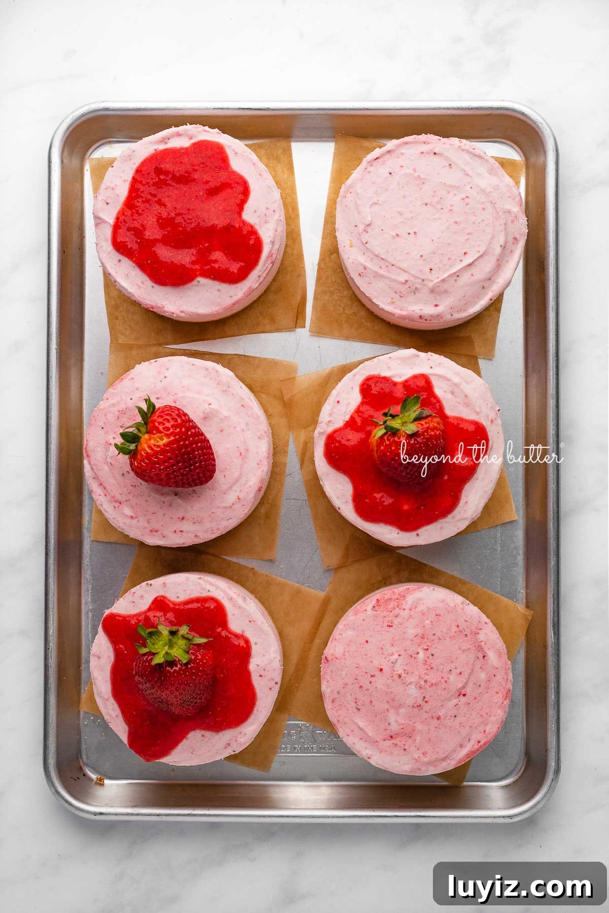Mini No-Bake Strawberry Cheesecakes (Individual Cups) 3 Individual no-bake strawberry cheesecakes topped with strawberry glaze and a fresh strawberry on a parchment paper-lined baking sheet | © Beyond the Butter®