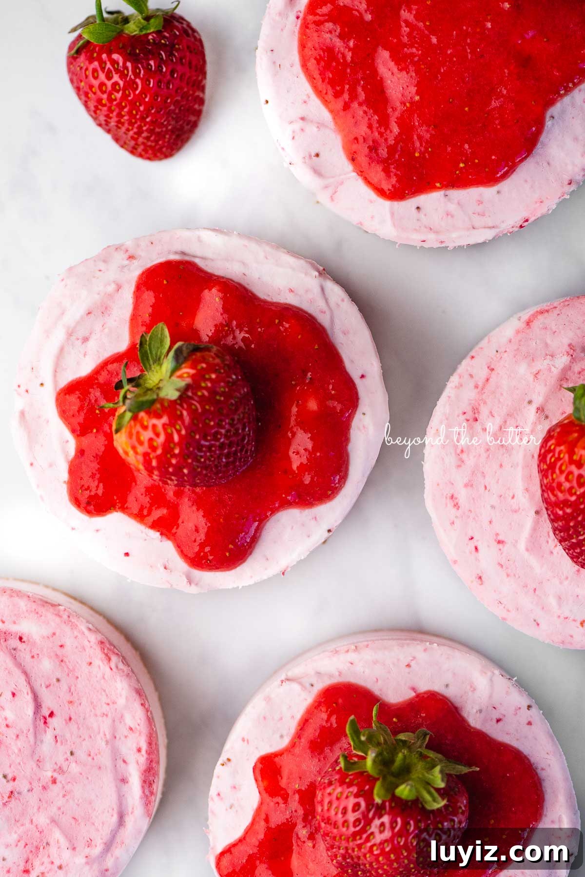 Mini No-Bake Strawberry Cheesecakes (Individual Cups) 5 Small 4-inch no-bake strawberry cheesecakes topped with strawberry glaze and a fresh strawberry on a parchment paper-lined baking sheet | © Beyond the Butter®