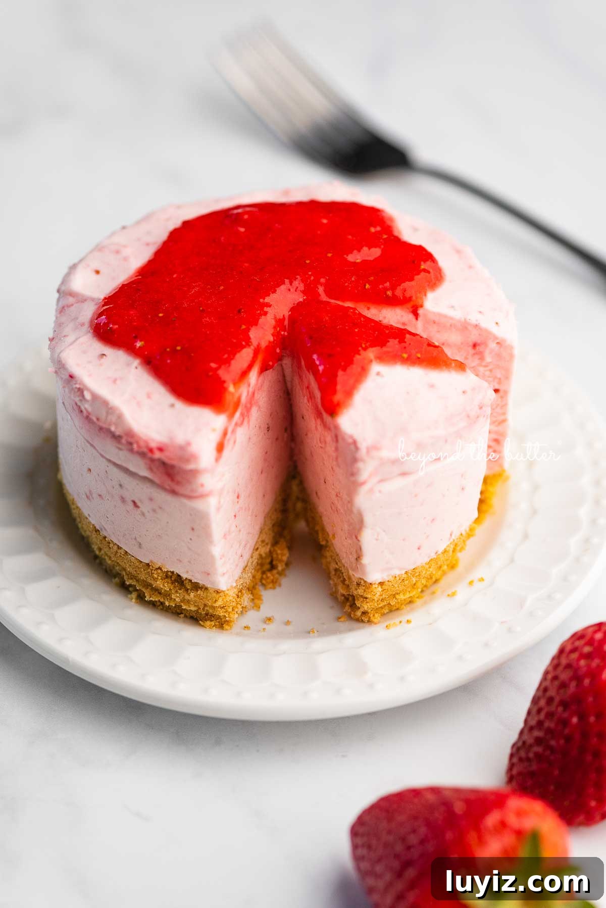 Mini No-Bake Strawberry Cheesecakes (Individual Cups) 6 Small 4-inch no-bake strawberry cheesecake on a white dessert plate with a fork and fresh strawberries nearby on a marbled background | © Beyond the Butter®