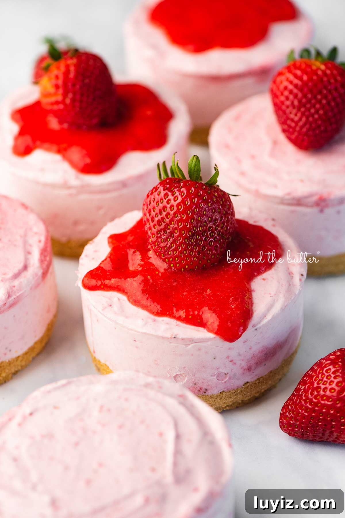 Mini No-Bake Strawberry Cheesecakes (Individual Cups) 7 Individual no-bake strawberry cheesecakes topped with strawberry glaze and a fresh strawberry on a parchment paper-lined baking sheet | © Beyond the Butter®