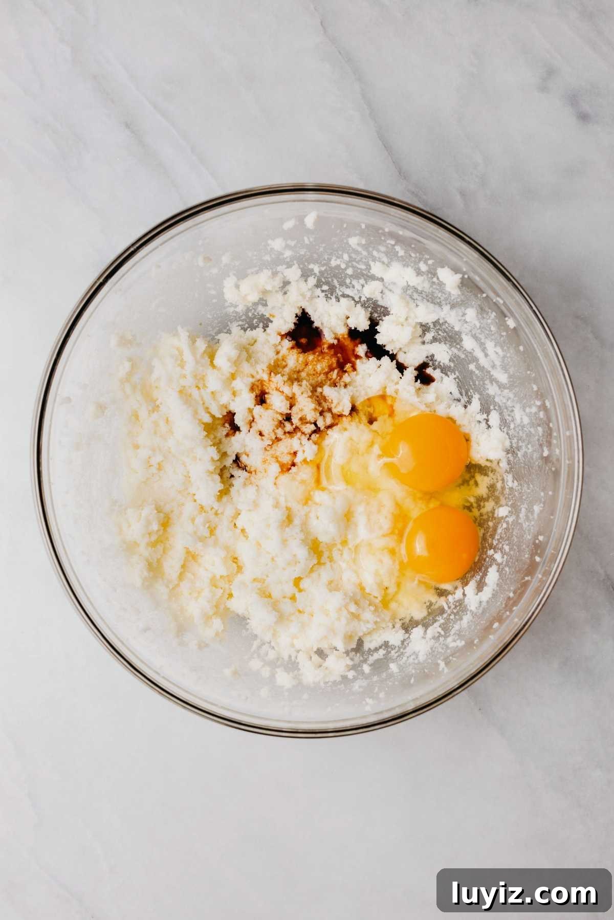 Classic Homemade Butter Cookies Recipe — Crisp, Melt-in-Your-Mouth Cookies 7 Unsalted butter and sugar blended together with eggs and vanilla extract added to the top in a glass bowl on a white and grey marbled background.