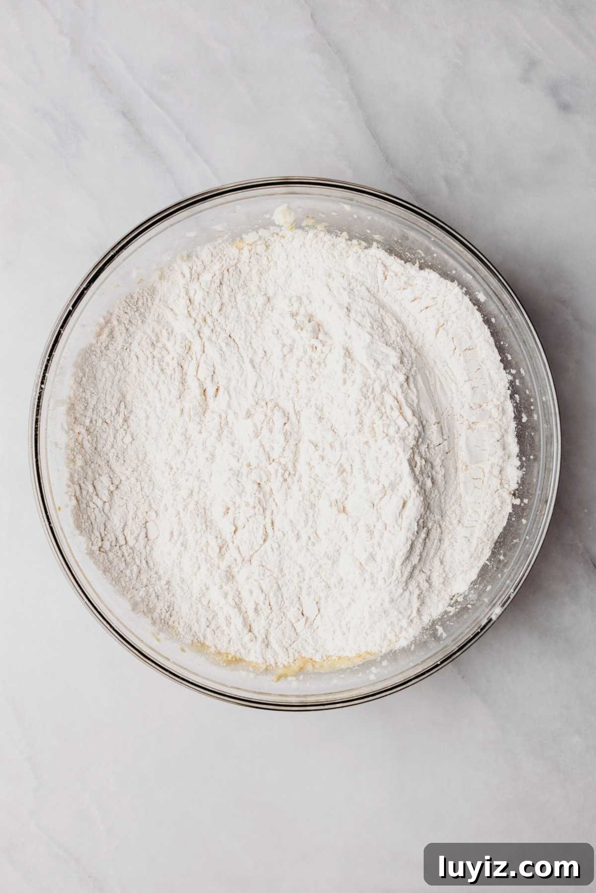 Classic Homemade Butter Cookies Recipe — Crisp, Melt-in-Your-Mouth Cookies 8 Dry ingredients added to the top of wet ingredients in a glass mixing bowl on a white and grey marbled background.