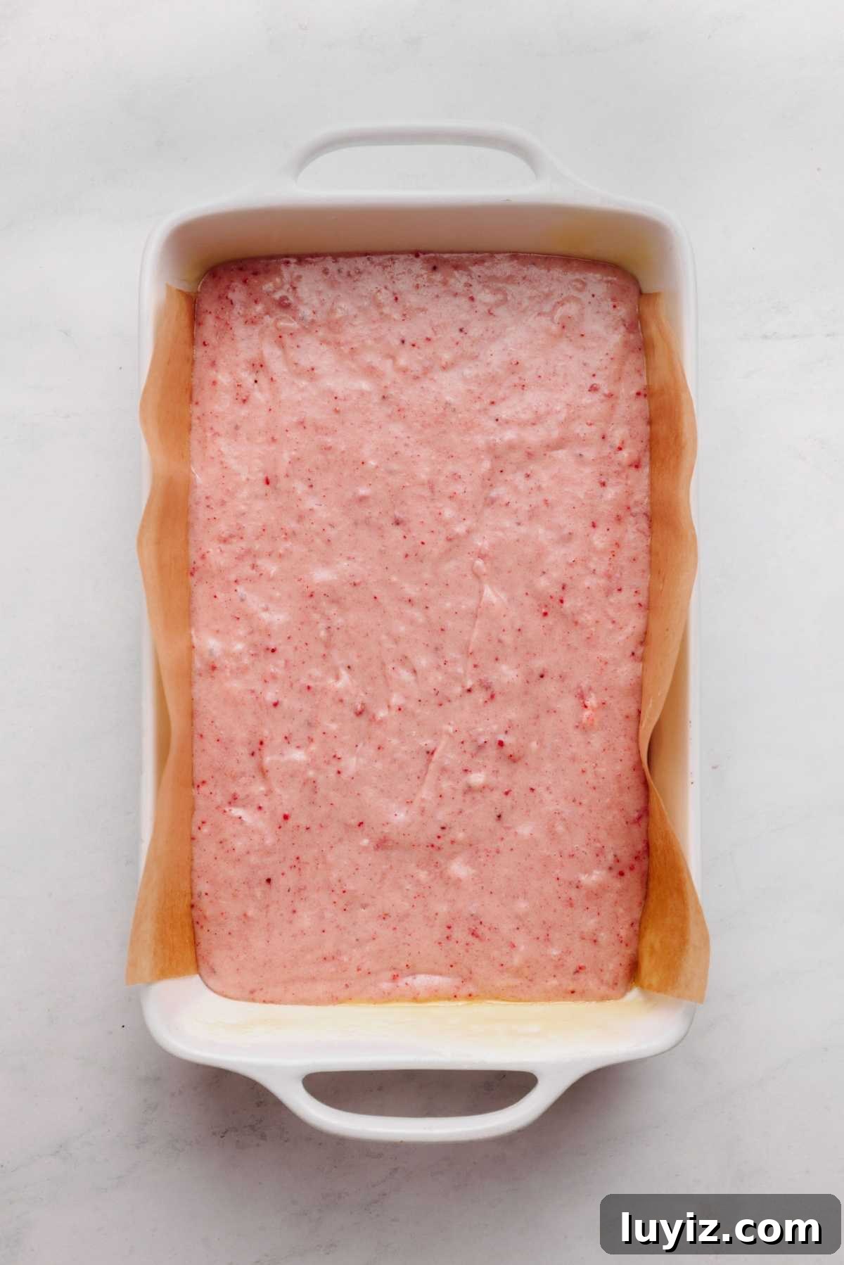 Mini Strawberry Sheet Cake Recipe: Quick Moist Fresh Strawberry Cake 14 Strawberry cake batter in a parchment paper lined ceramic baking tin on a white and grey textured background.