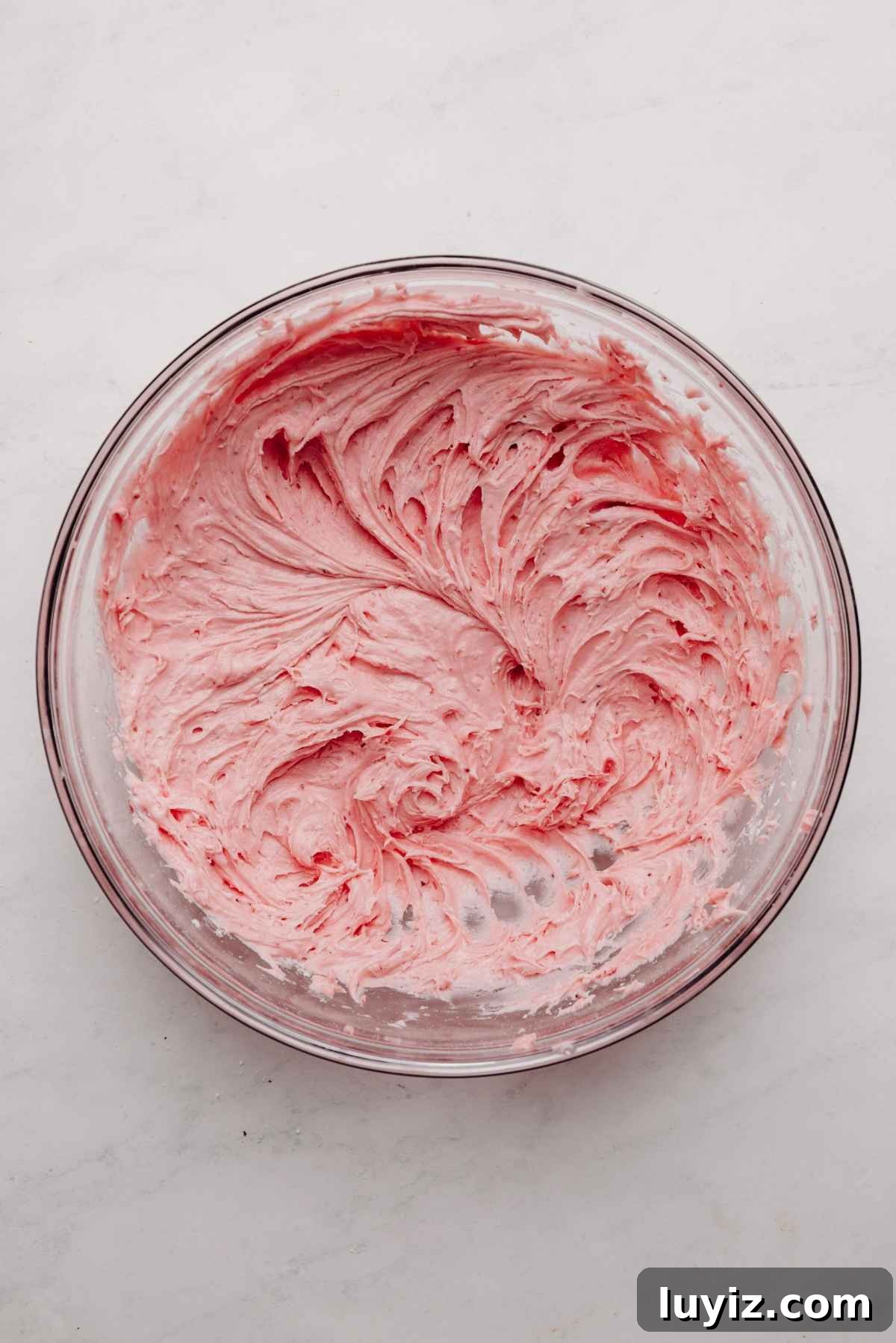 Mini Strawberry Sheet Cake Recipe: Quick Moist Fresh Strawberry Cake 18 Strawberry buttercream frosting in a large glass mixing bowl on a white and grey textured background.