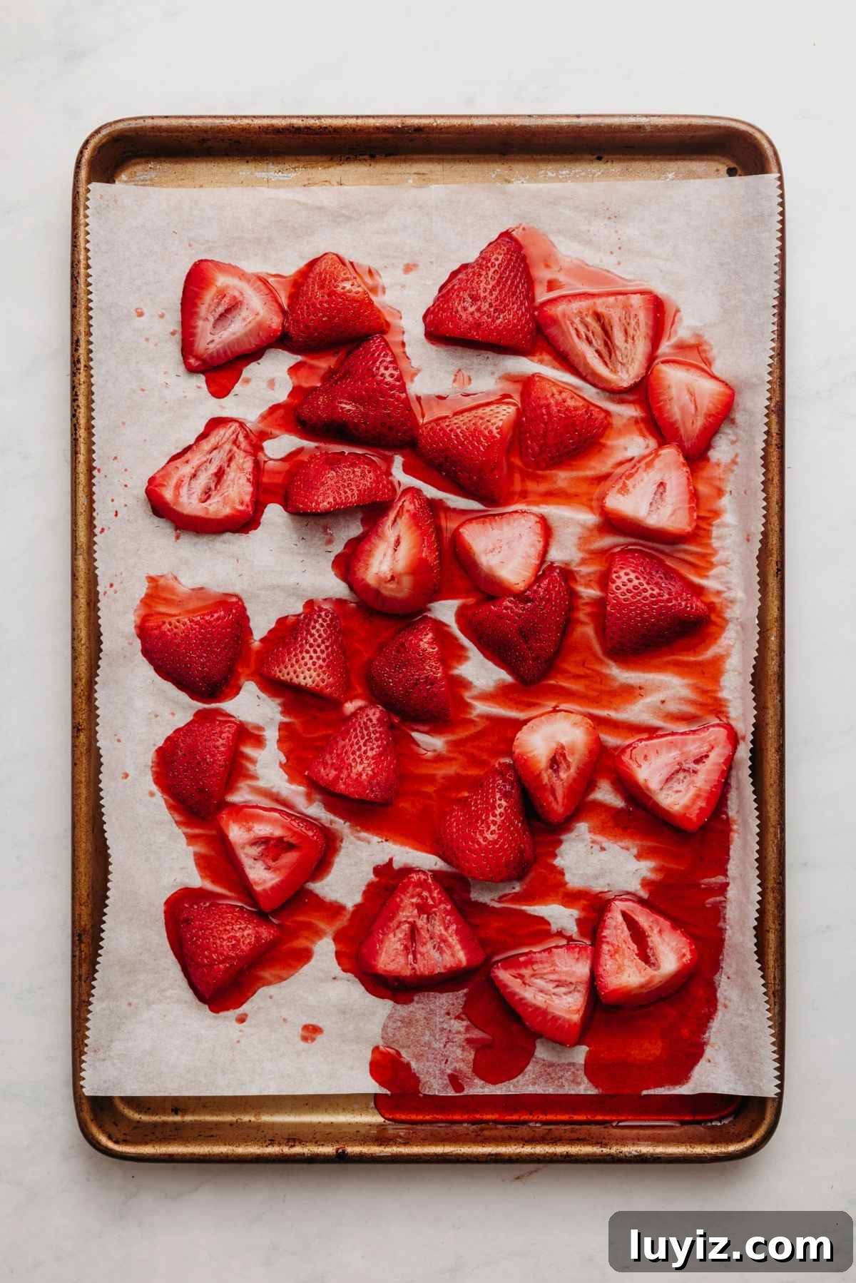 Mini Strawberry Sheet Cake Recipe: Quick Moist Fresh Strawberry Cake 7 Roasted strawberries with the juices on a parchment paper lined baking sheet on a white and grey textured background.