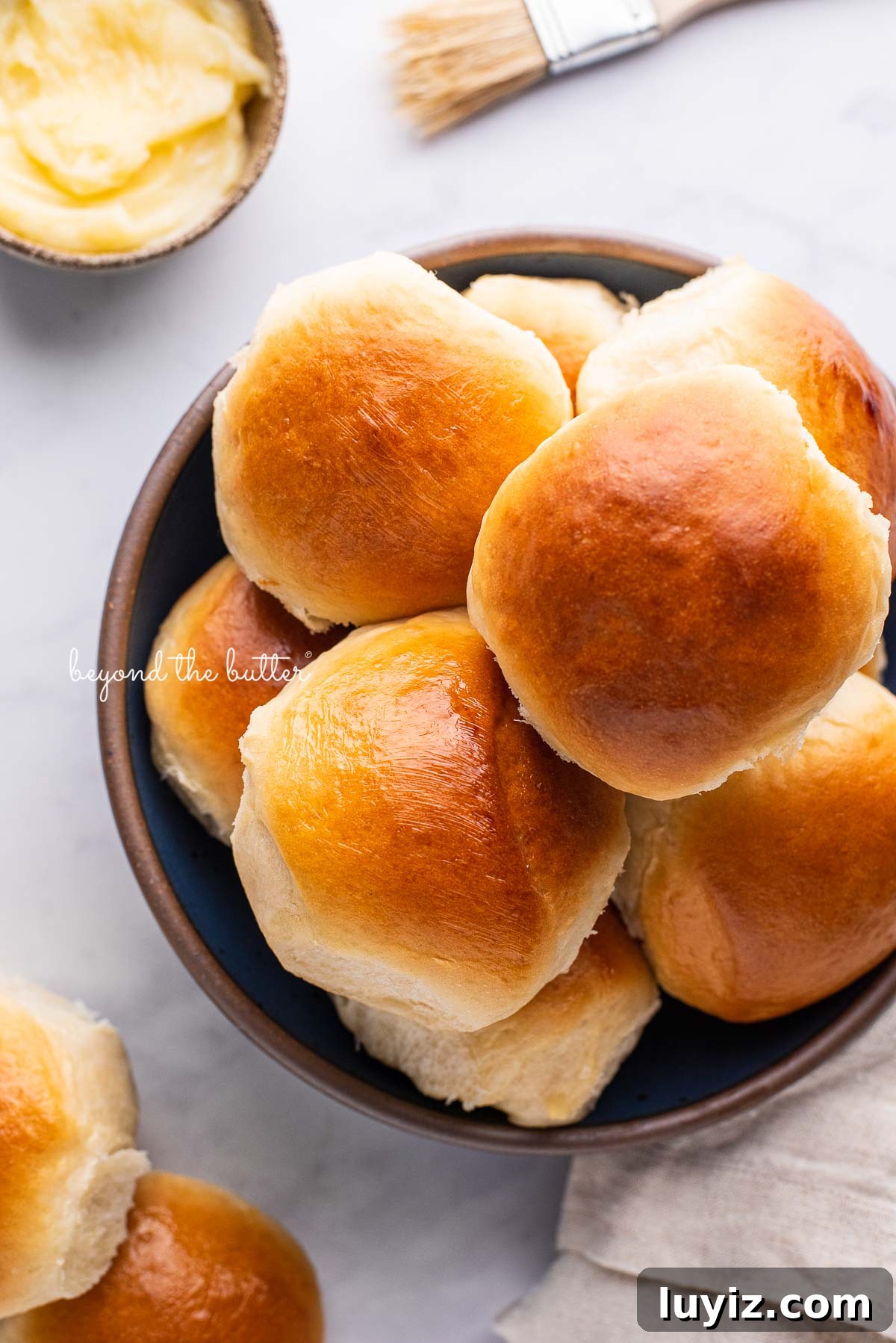 Soft Honey Butter Dinner Rolls Recipe for Fluffy, Golden Buns 3 Blue ceramic bowl filled with homemade honey butter dinner rolls with a small bowl of honey butter and brush next to it on a white marbled background | © Beyond the Butter®