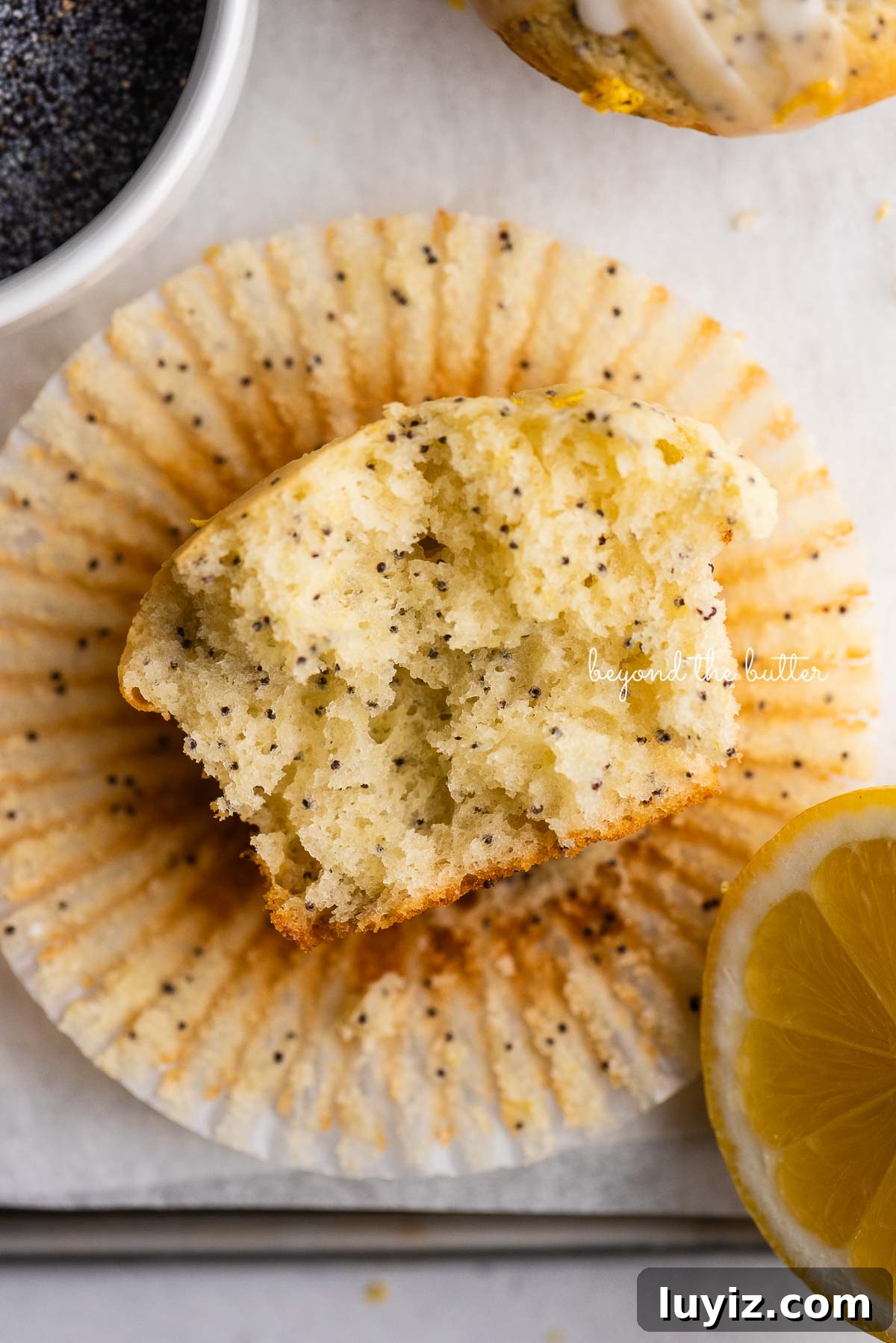 Lemon Poppy Seed Muffins with Zesty Glaze Recipe 12 Half eaten lemon poppyseed muffin on top of muffin liner on white background with bowl of poppyseeds and slice of lemon.