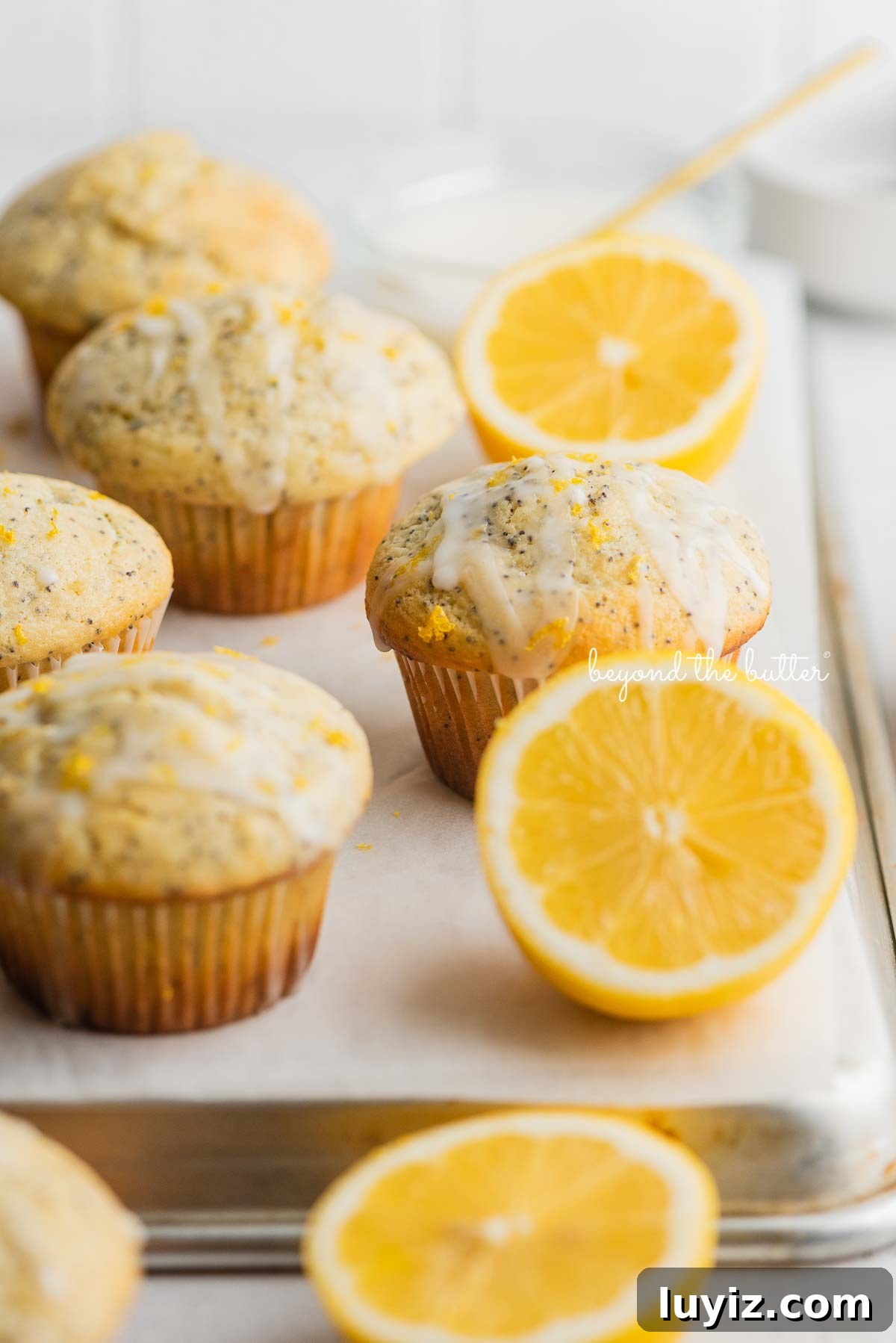 Lemon Poppy Seed Muffins with Zesty Glaze Recipe 13 Lemon poppyseed muffins randomly placed on a white parchment paper inverted baking sheet with bowl of glaze and half slices of lemons next to them.
