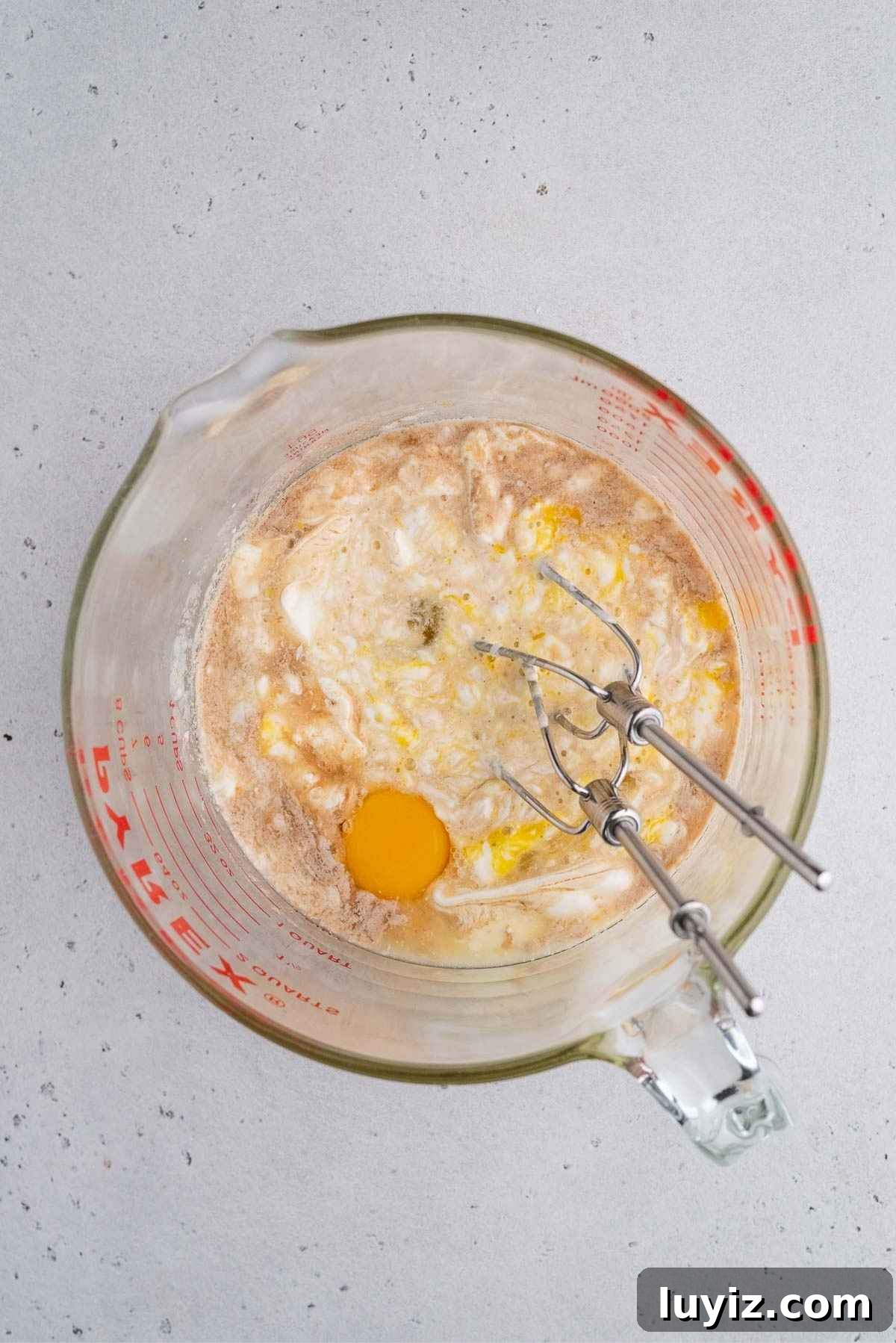 Lemon Poppy Seed Muffins with Zesty Glaze Recipe 6 Mixing the lemon poppy seed muffin wet ingredients together in a large glass measuring bowl with hand mixer whisks resting in bowl on a light grey background.