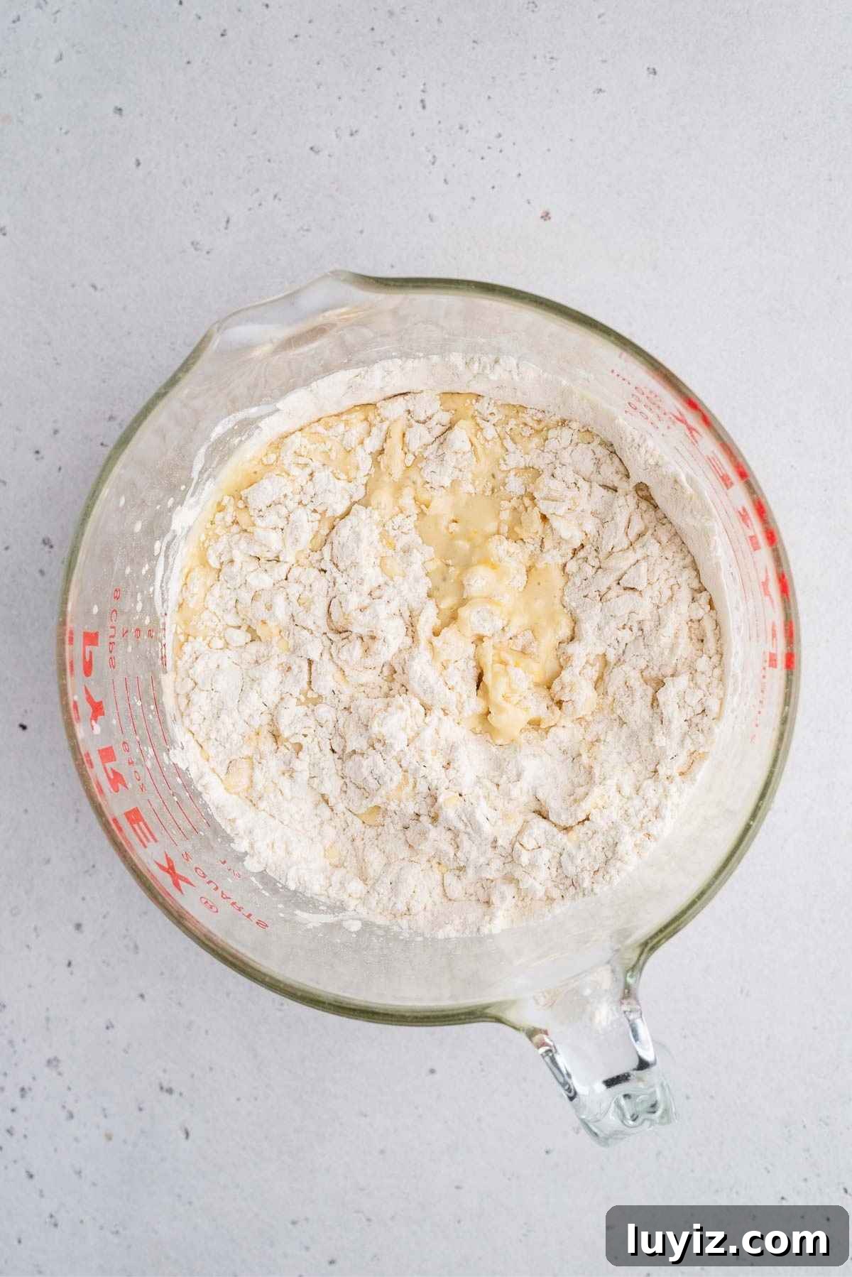 Lemon Poppy Seed Muffins with Zesty Glaze Recipe 7 Lemon poppy seed muffin batter in a large glass measuring bowl with dry ingredients partially mixed in.