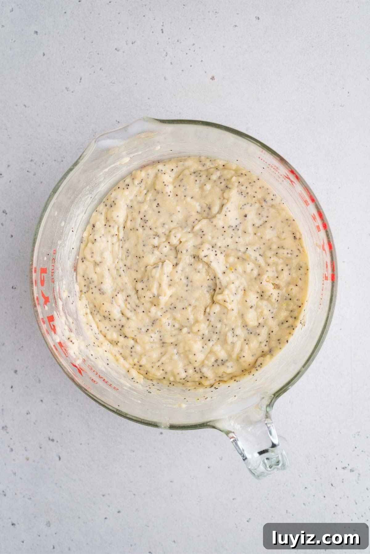 Lemon Poppy Seed Muffins with Zesty Glaze Recipe 8 Melted butter and poppy seeds stirred into the lemon muffin batter in a large glass measuring bowl on a light grey background.