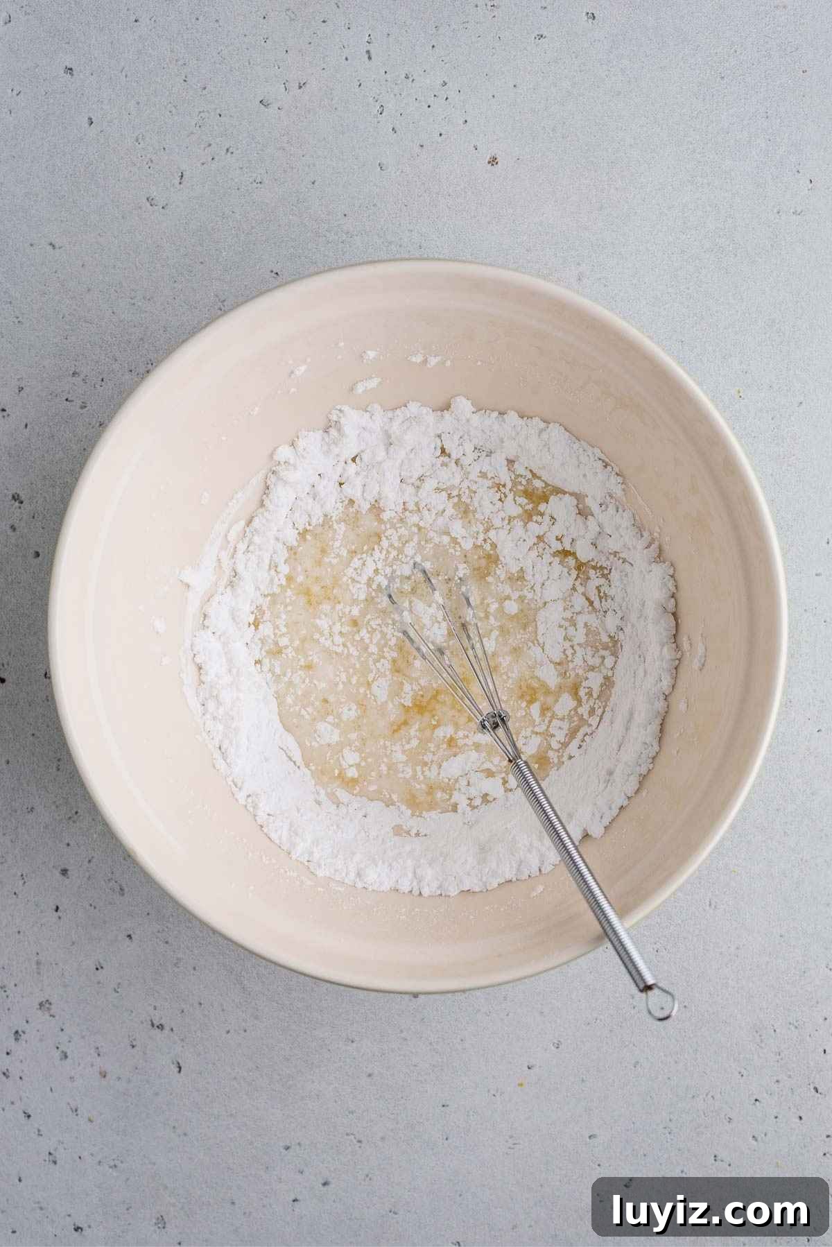 Lemon Poppy Seed Muffins with Zesty Glaze Recipe 10 Whisking the lemon glaze together in a small ceramic bowl and a mini whisk on a light grey background.