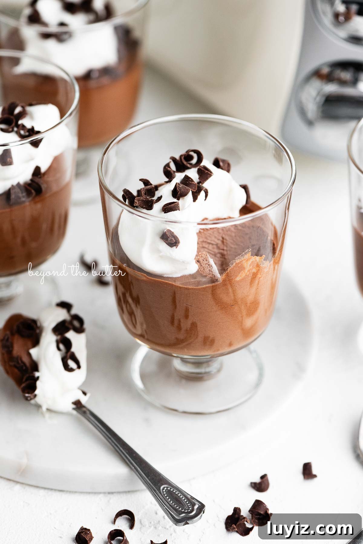 Blender Chocolate Mousse Recipe — Creamy No-Bake Dessert 2 Parfait goblets filled with chocolate mousse, whipped cream, and chocolate curls with a spoonful laying next to one on a white marble slab and white textured background.