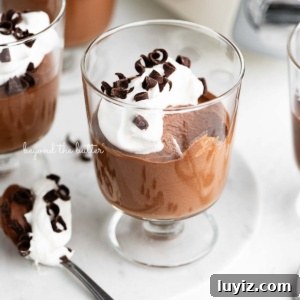 Blender Chocolate Mousse Recipe — Creamy No-Bake Dessert 8 Parfait goblets filled with chocolate mousse, whipped cream, and chocolate curls with a spoonful laying next to one on a white marble slab and white textured background.