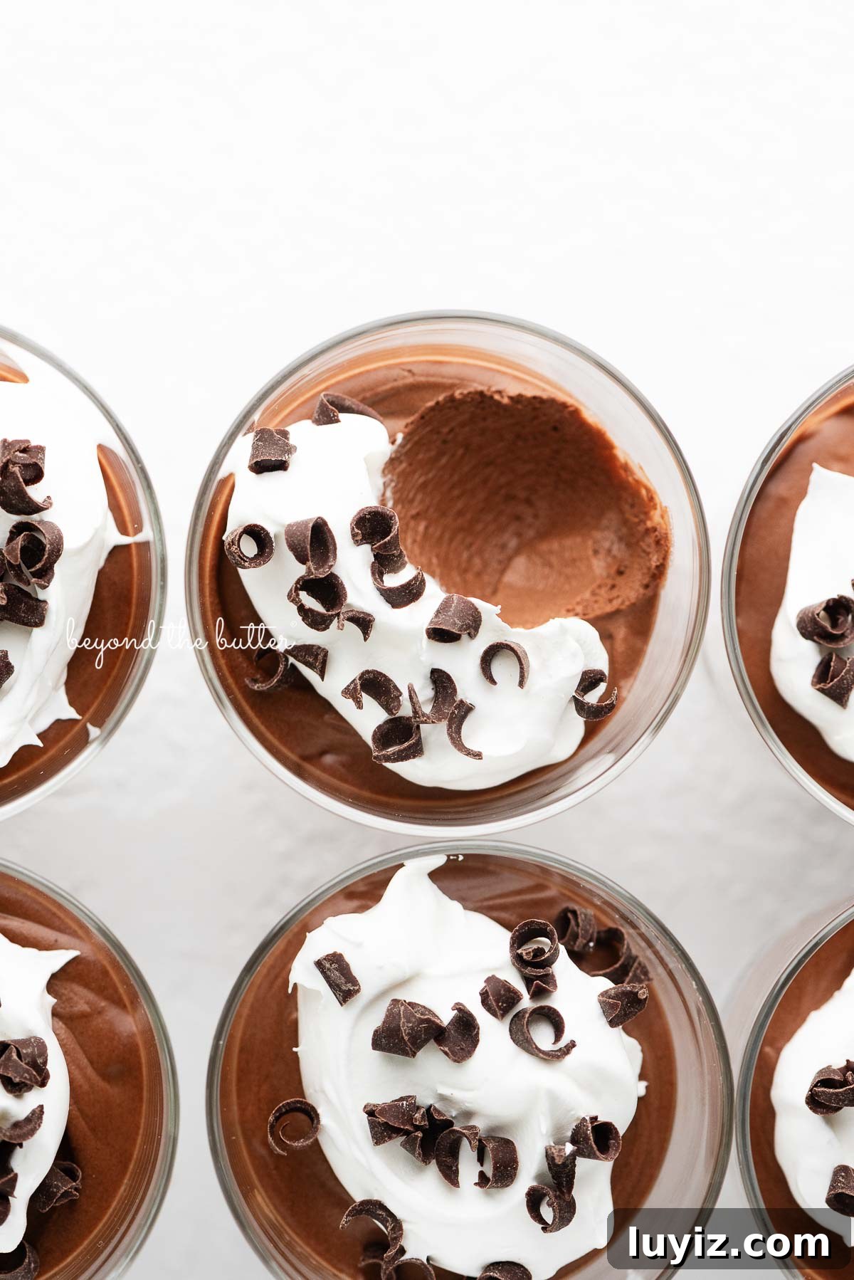 Blender Chocolate Mousse Recipe — Creamy No-Bake Dessert 7 Six parfait goblets filled with chocolate mousse, whipped cream, and chocolate curls with a spoonful removed from the top center goblet on a white textured background.