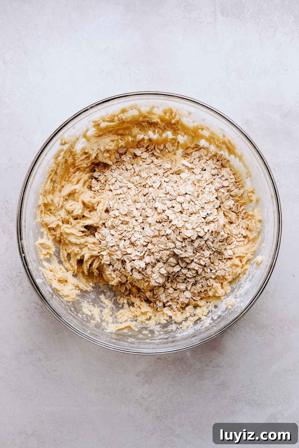 Salted Caramel Oatmeal Chocolate Chip Cookies 7 Rolled oats added to the top of oatmeal cookie dough mixture in a glass mixing bowl on a white grey marbled background.