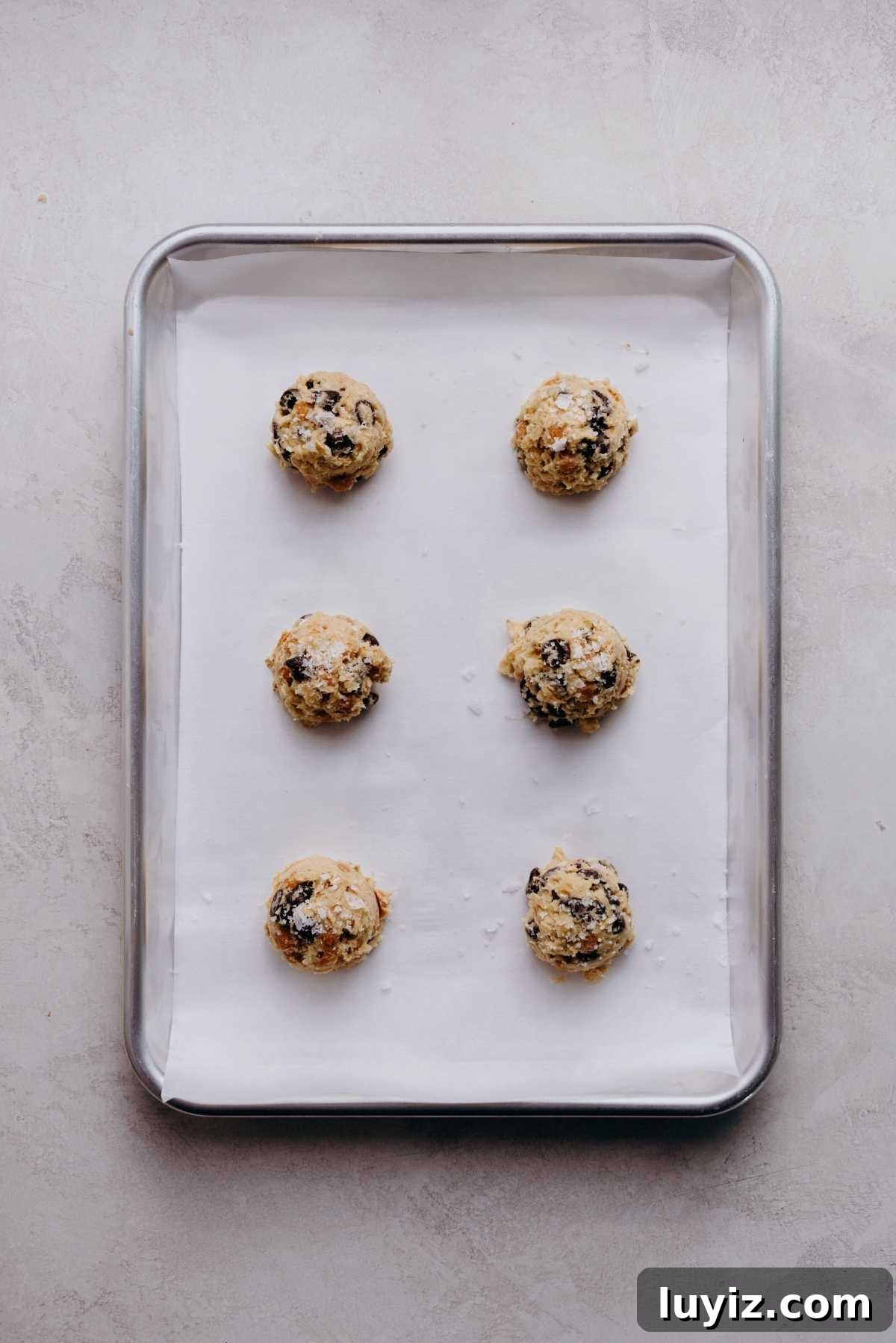Salted Caramel Oatmeal Chocolate Chip Cookies 9 Scoops of oatmeal chocolate chip salted caramel cookie dough topped with flaky sea salt on a white parchment paper lined cookie sheet.